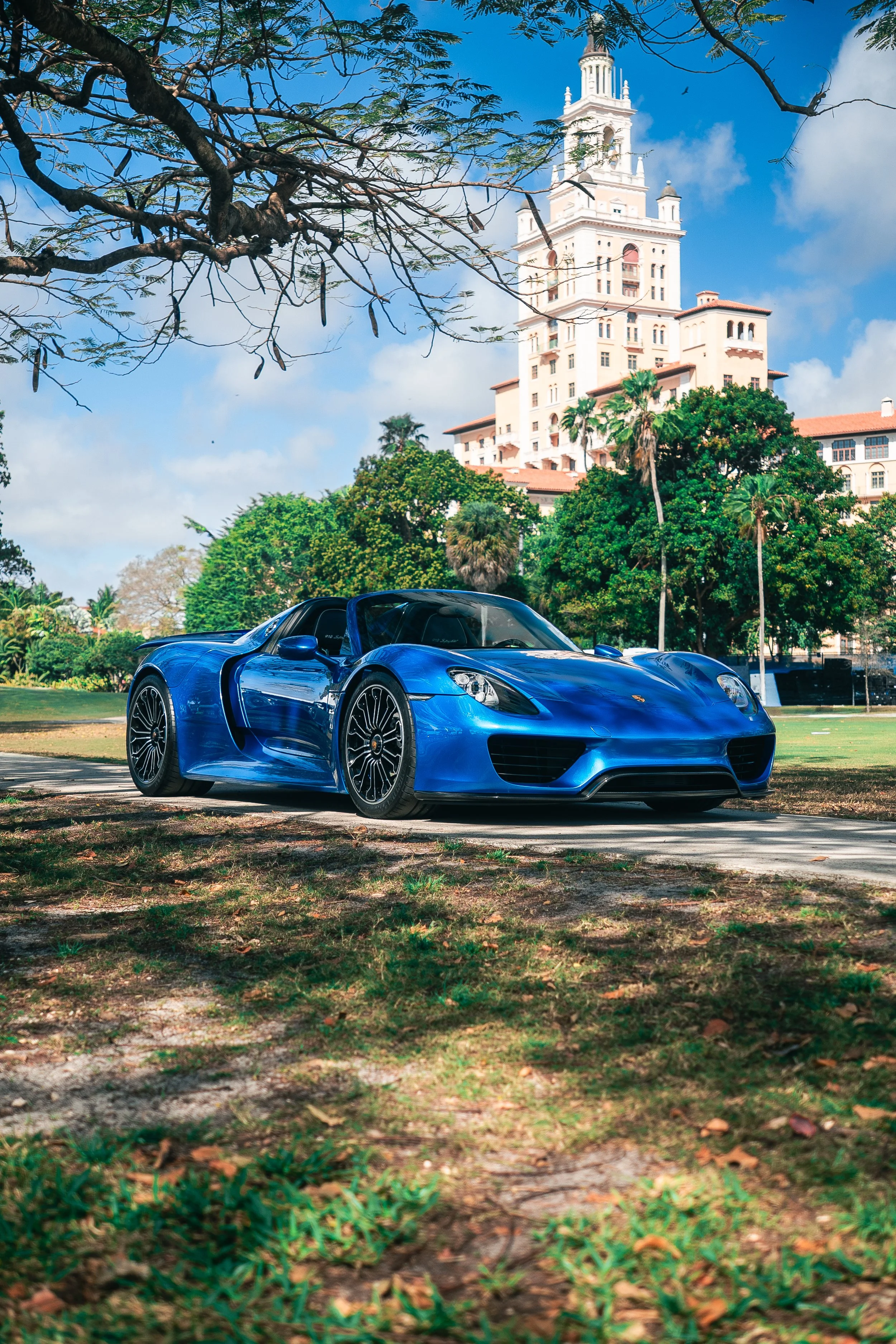 A blue Porsche 918 Spyder parked on a grassy area with trees and a large historic building with a tower in the background under a partly cloudy sky.
