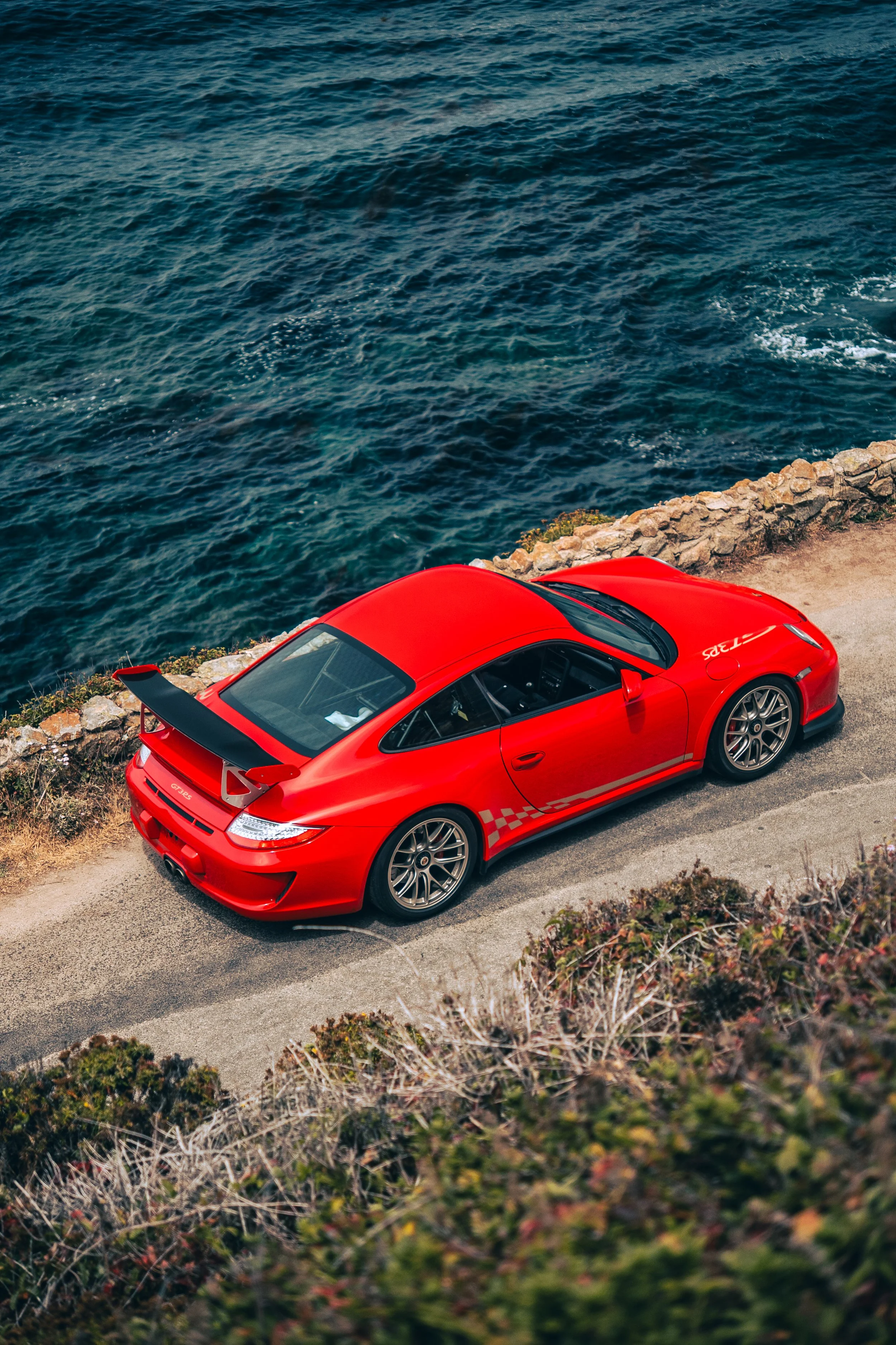 Red Porsche 911 GT3RS 997 parked near a rocky coastline with water in the background.