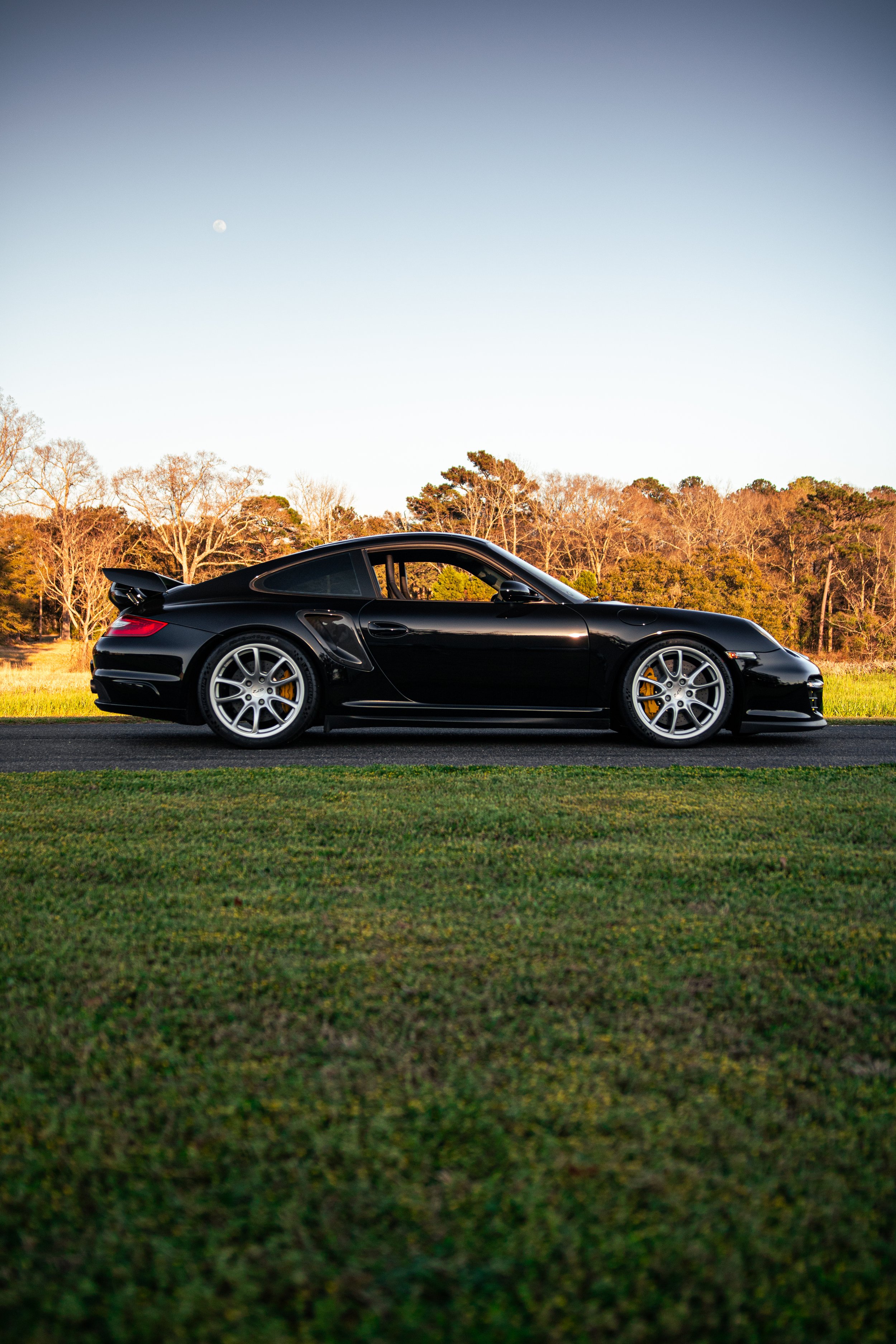 A black Porsche 911 GT2 997 parked on grass with trees in the background and the moon visible in the sky.