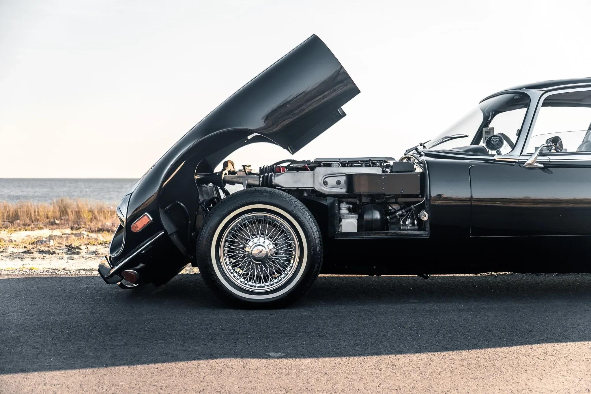 A black Jaguar E-Type Coupe XKE with its hood open, showing the engine, parked on a road by the beach with grass and water in the background.