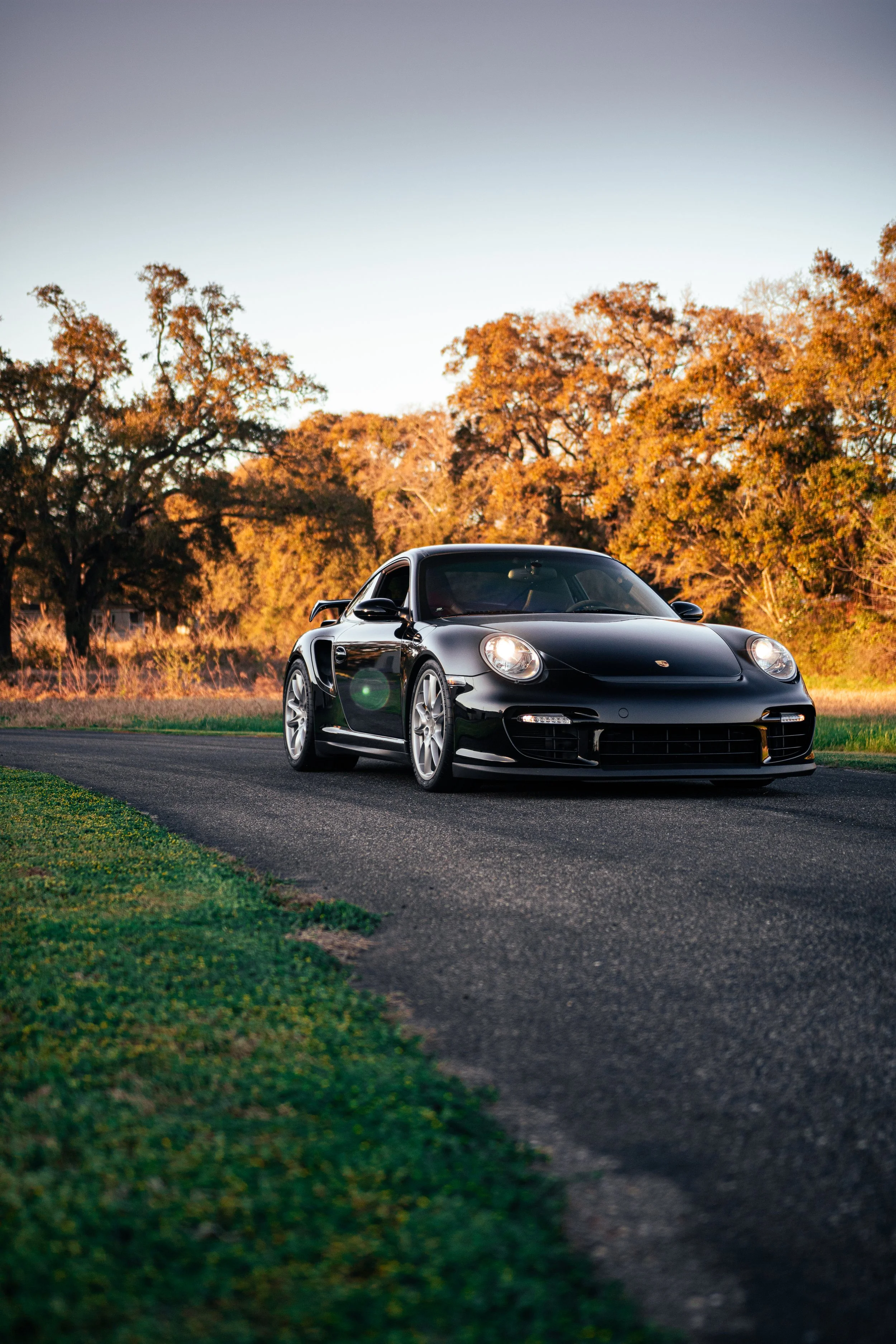 A black Porsche 911 GT2 997 parked on a rural road during sunset, with trees in autumn colors in the background.