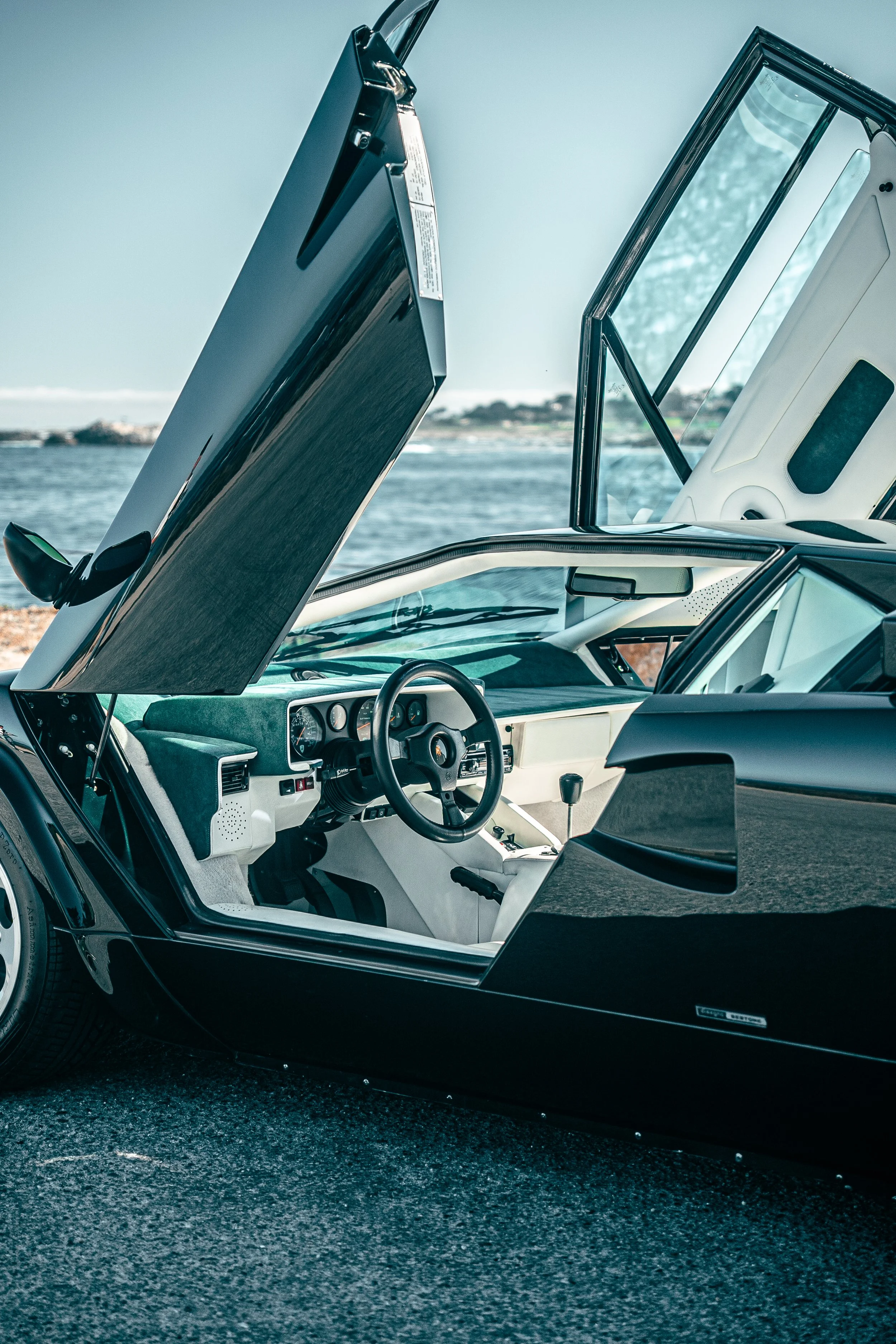 Interior of a Lamborghini Countach with its doors open, showing the dashboard and steering wheel, parked near a body of water with rocks and trees in the background.