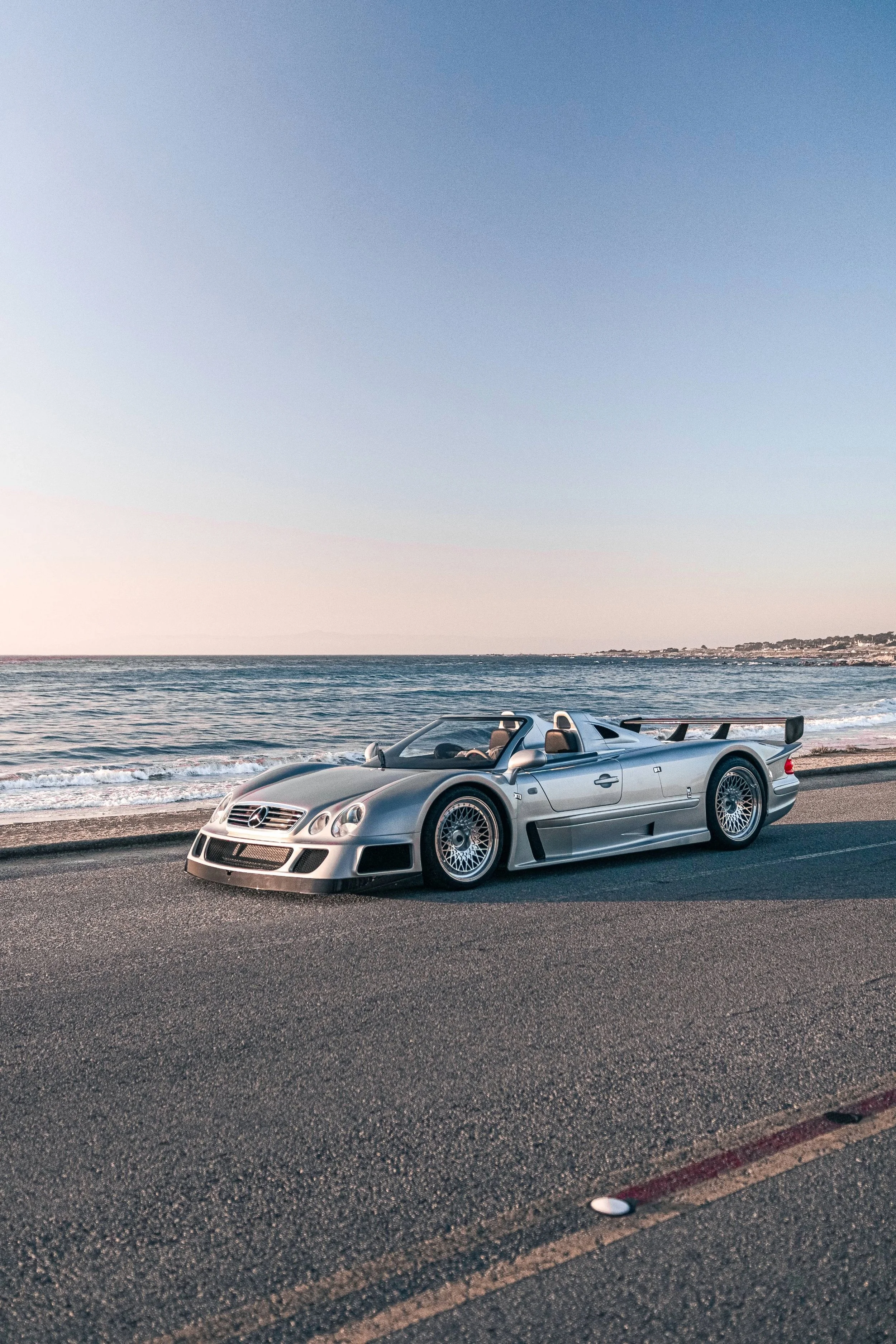 A silver Mercedes-Benz CLK GTR Roadster parked on a coastal road with the ocean and a distant shoreline in the background during sunset.