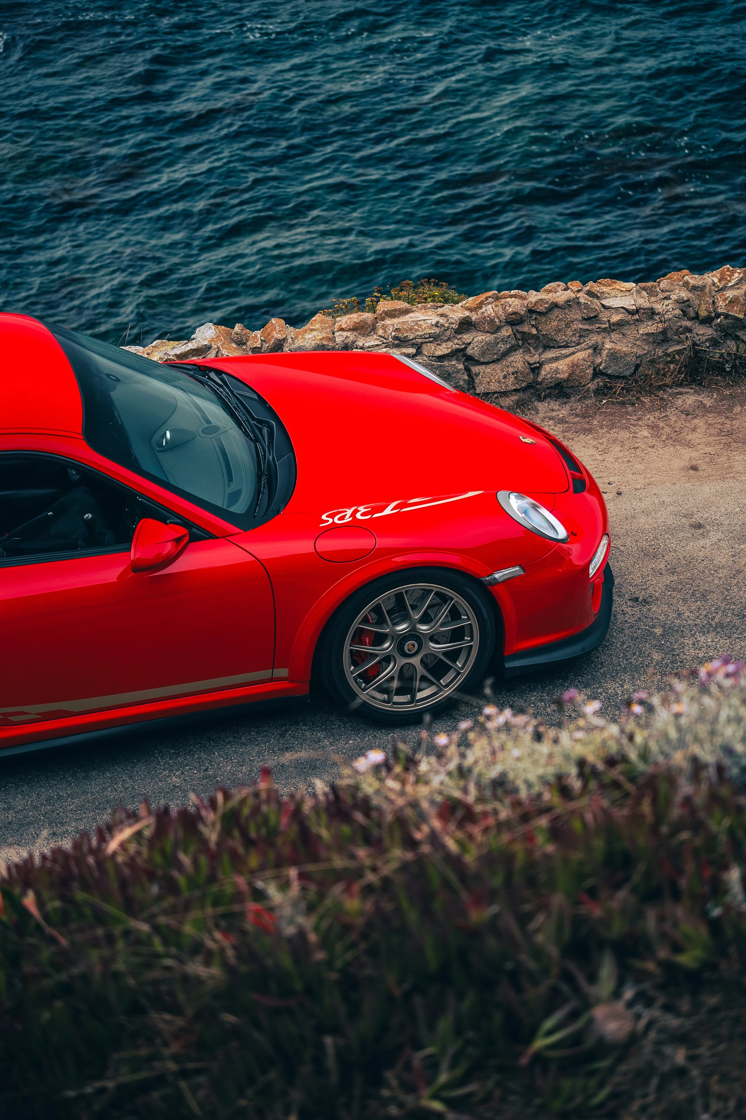 Red Porsche 911 GT3RS 997 parked beside a stone wall overlooking the ocean.