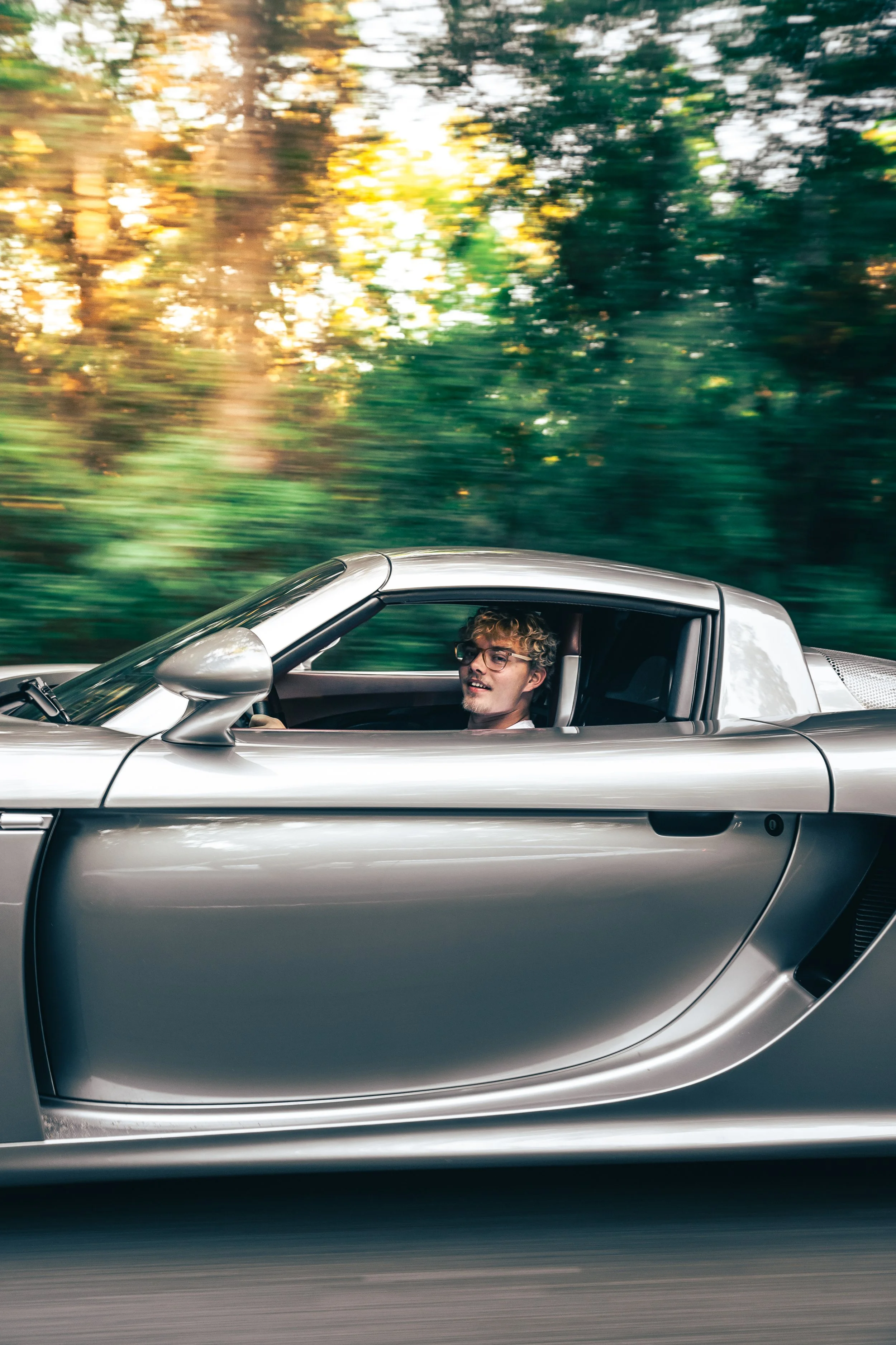 A man with curly hair and glasses driving a silver Porsche Carrera GT on a road surrounded by trees with blurred leaves, suggesting motion.