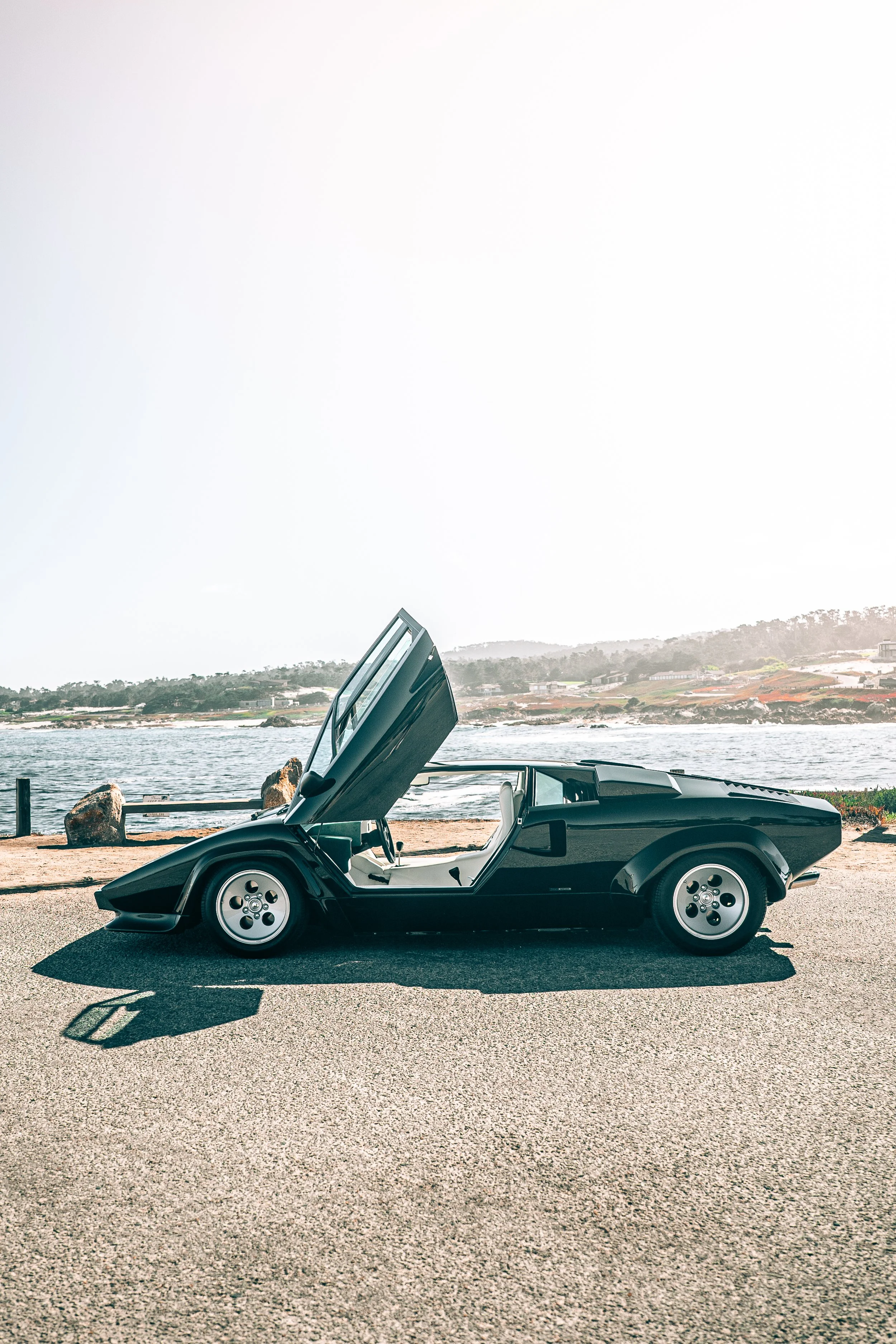 A Lamborghini Countach with its doors open parked near a body of water with rocky shoreline and hills in the background.