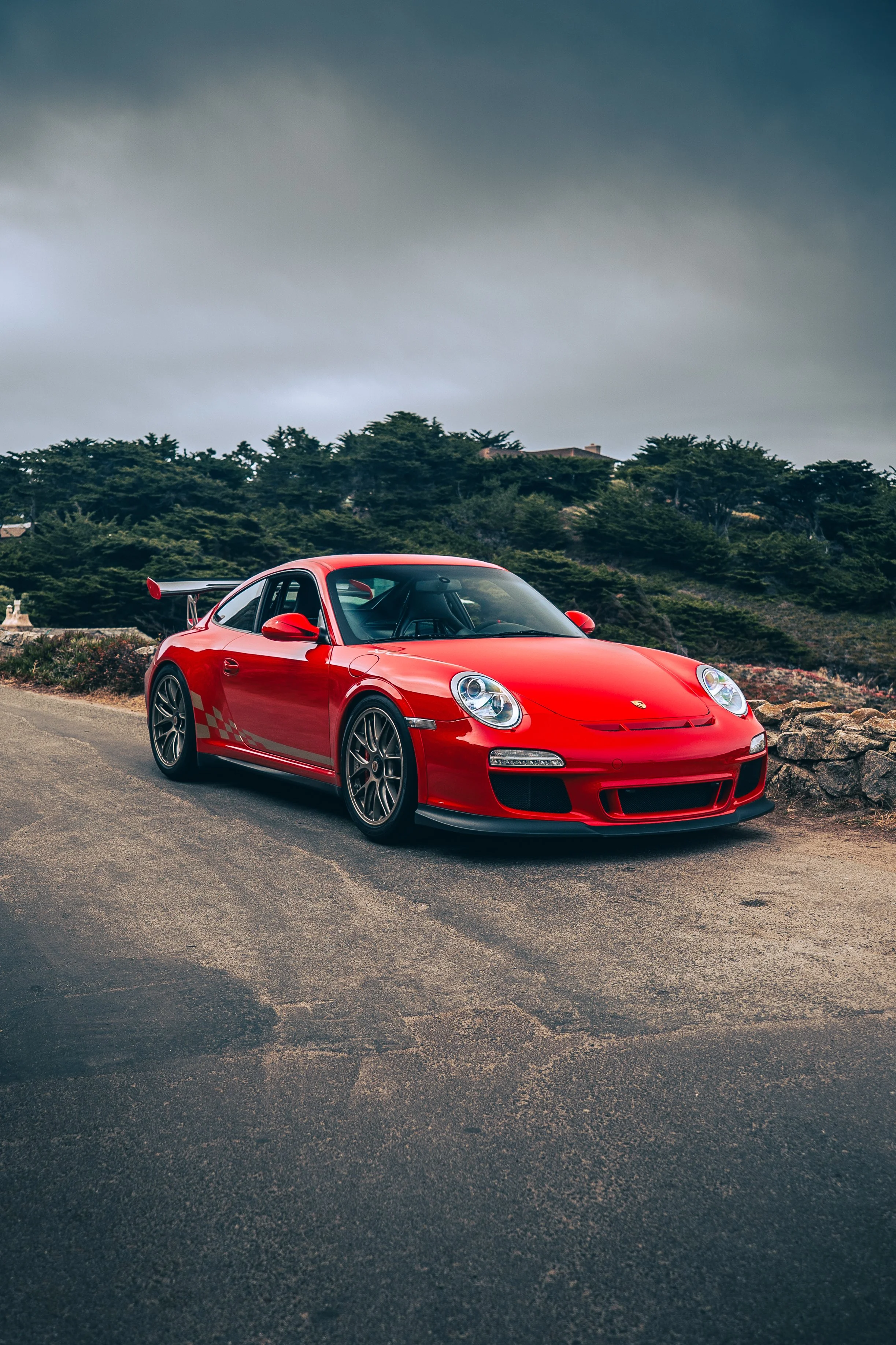 Red Porsche 911 GT3RS 997 parked on the side of the road with trees and cloudy sky in the background.
