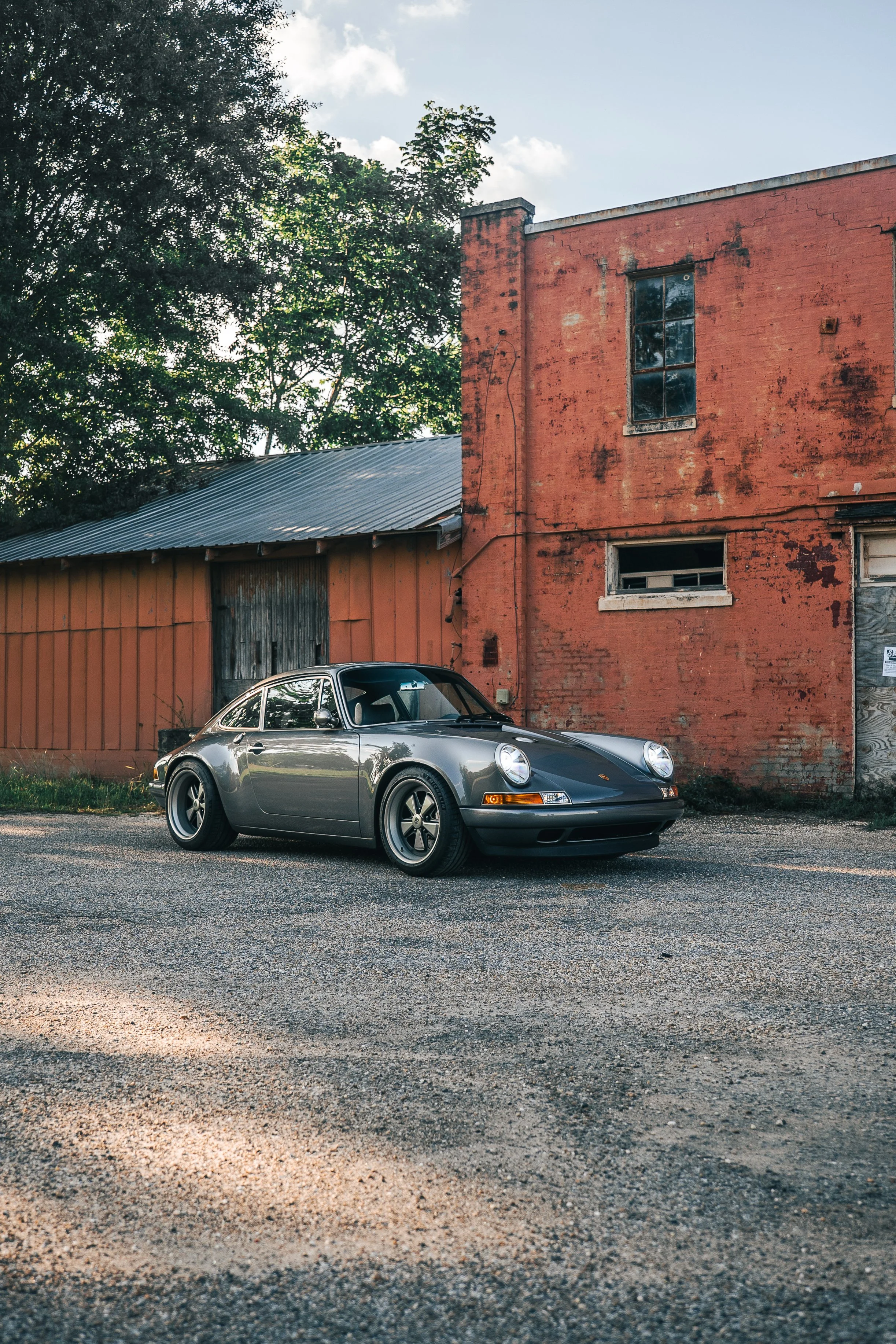 A gray Singer Porsche 911 is parked on a gravel lot in front of an old, weathered red brick building with a metal roof and windows. There are trees in the background and the sky is partly cloudy.
