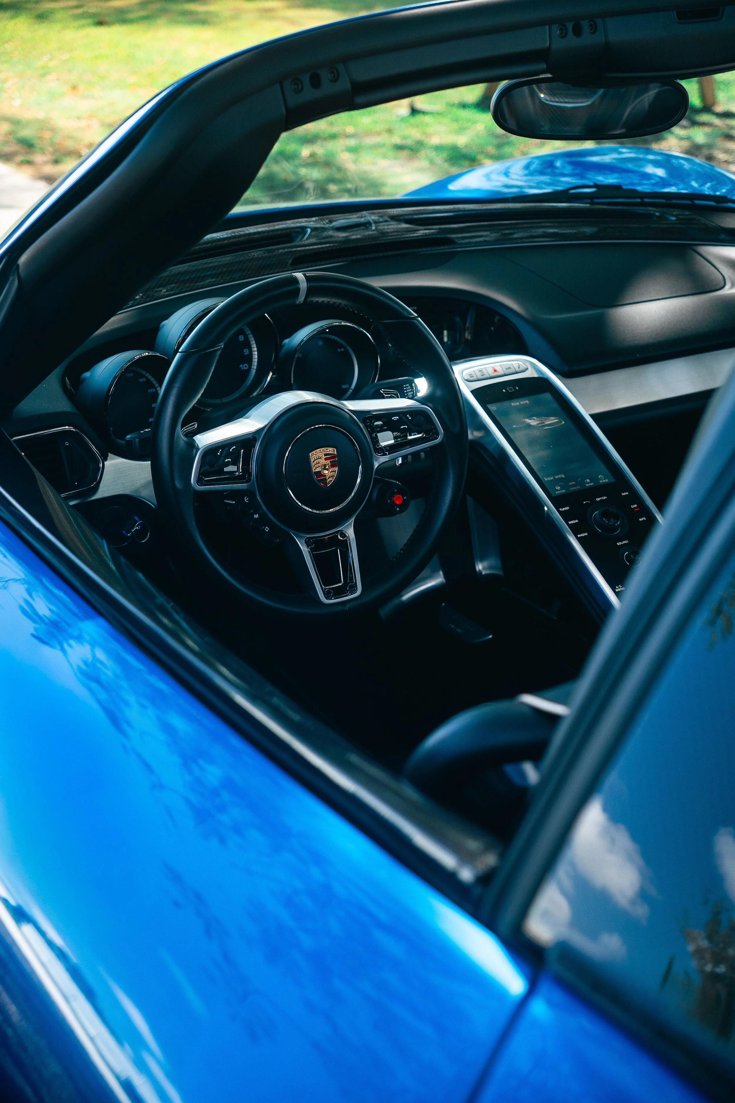 Interior of a blue Porsche 918 Spyder, showing the steering wheel with Porsche logo, dashboard, and a large touchscreen display, viewed through the driver's side window.