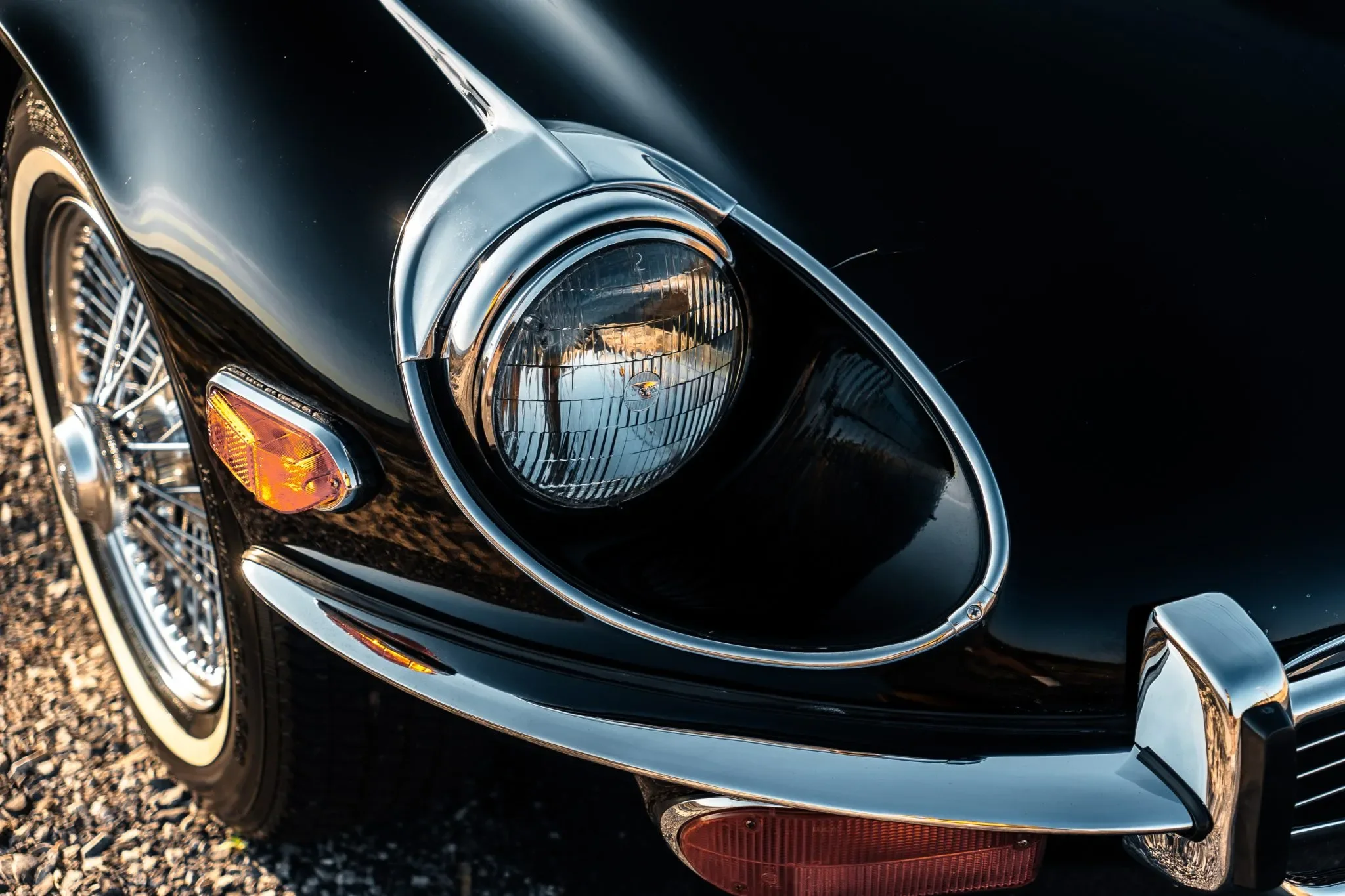 Close-up of a black Jaguar E-Type Coupe XKE front headlight, chrome detailing, turn signal, and wire-spoked wheel.