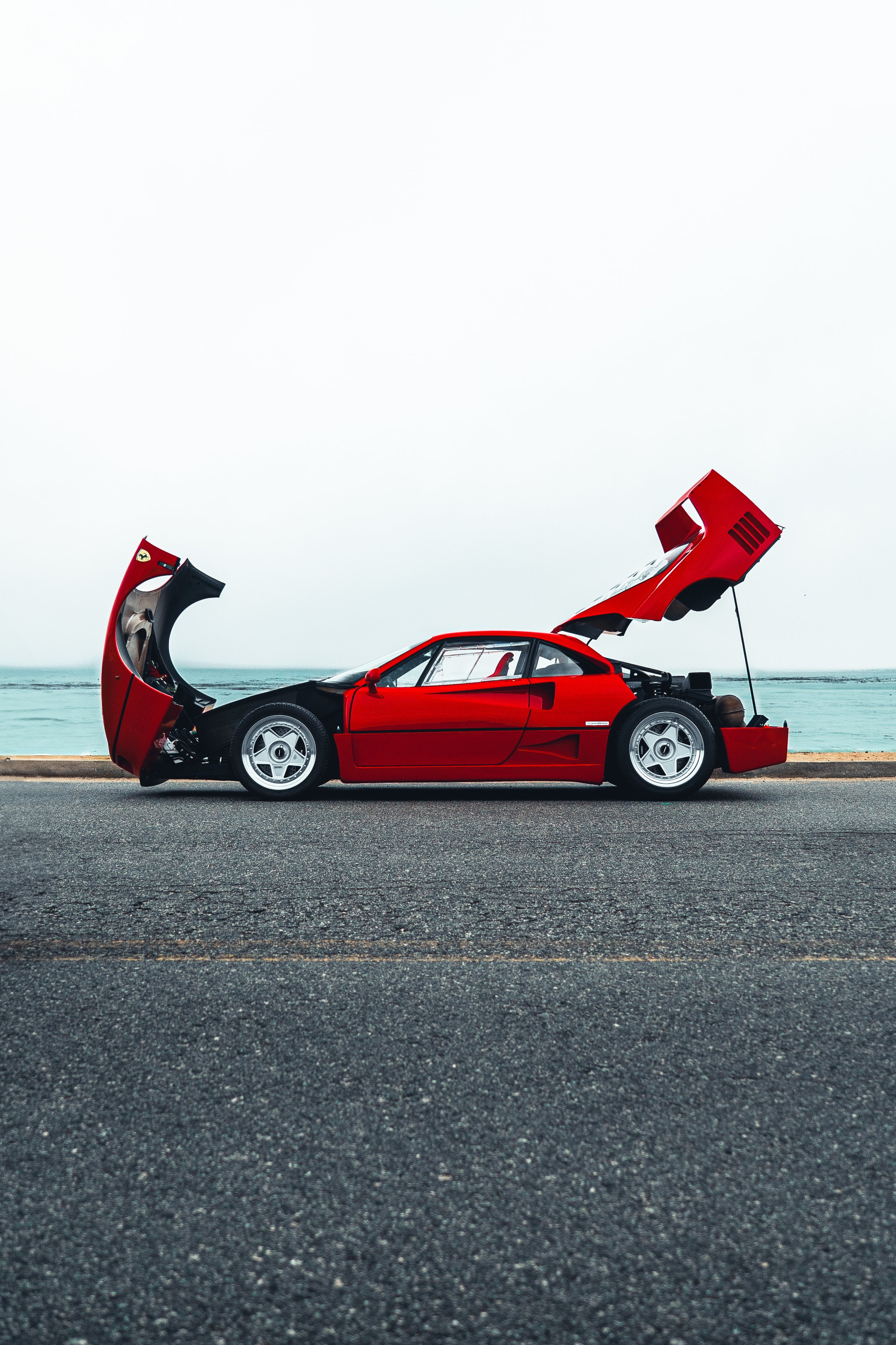 A Ferrari F40 is parked by the beach with its front and rear hoods open, with the ocean in the background under a cloudy sky.