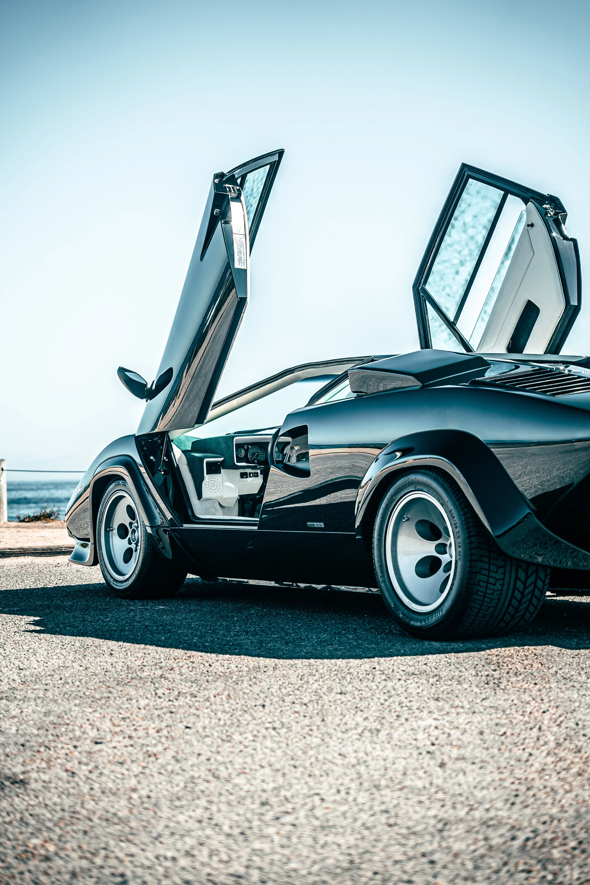 A green Lamborghini Countach with doors open, parked on a gravel surface by the ocean, with a clear sky in the background.