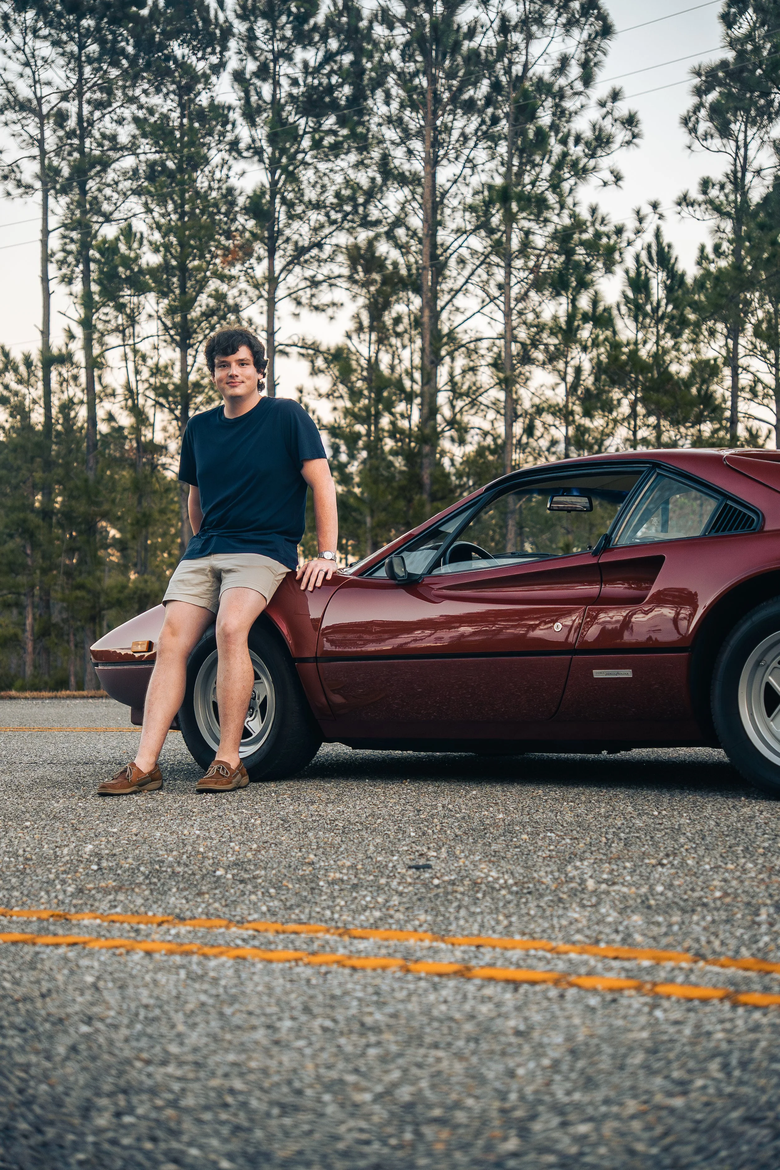 A young man wearing a navy blue t-shirt, beige shorts, and brown shoes leaning against a Ferrari 308 GTB parked on a road with trees in the background.