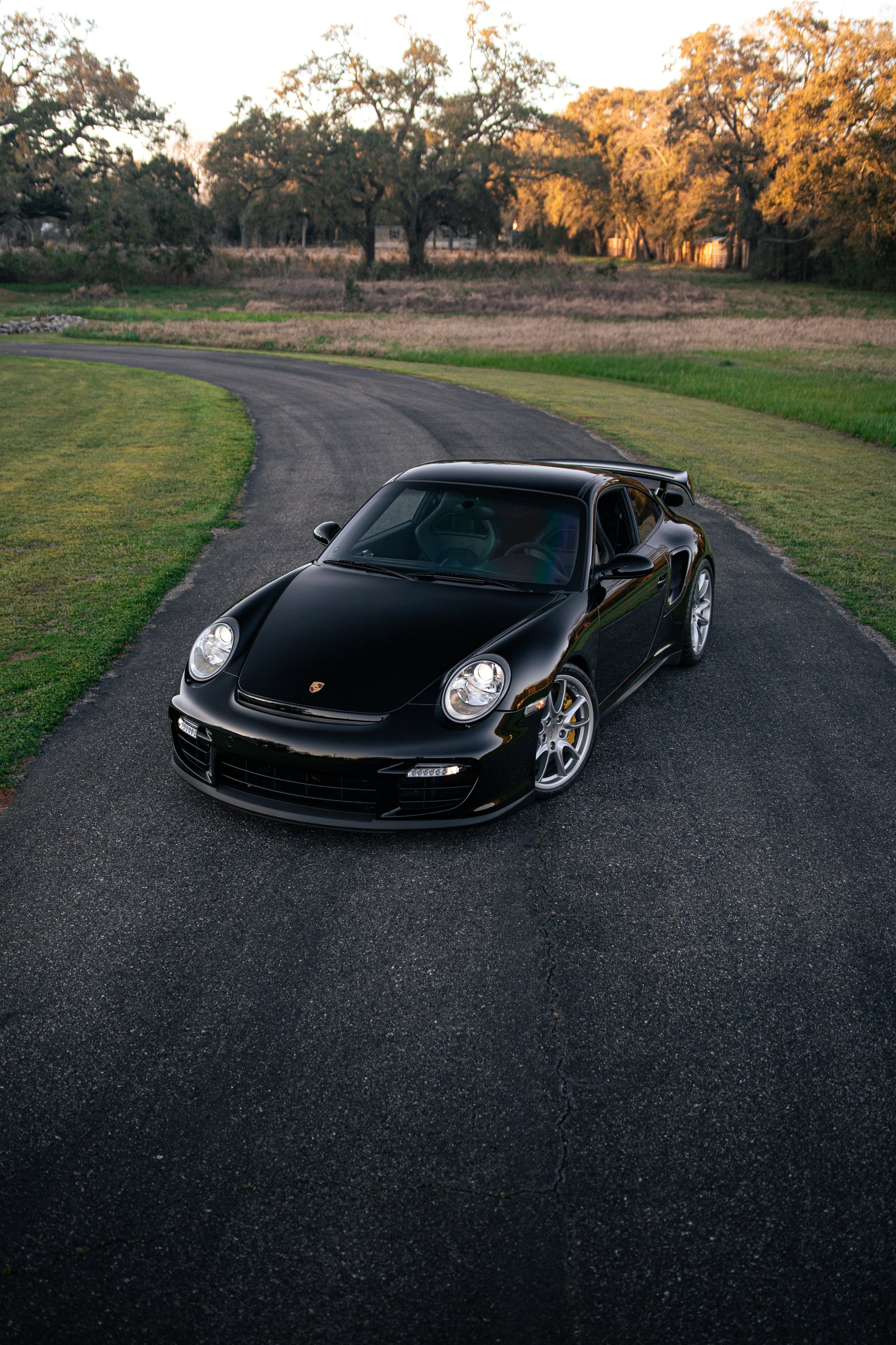 A black Porsche 911 GT2 997 parked on a curved asphalt driveway with greenery on the sides and trees in the background at sunset.