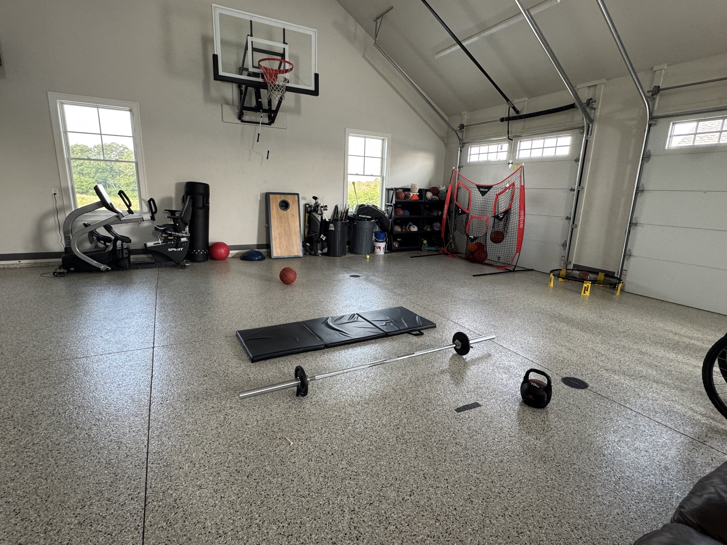 A spacious garage converted workout area with gym equipment, basketball hoop, and sports gear. Includes weightlifting barbell, exercise bike, a small basketball hoop, a basketball storage rack, and a protective padded mat on the floor. Two windows bring in natural light.