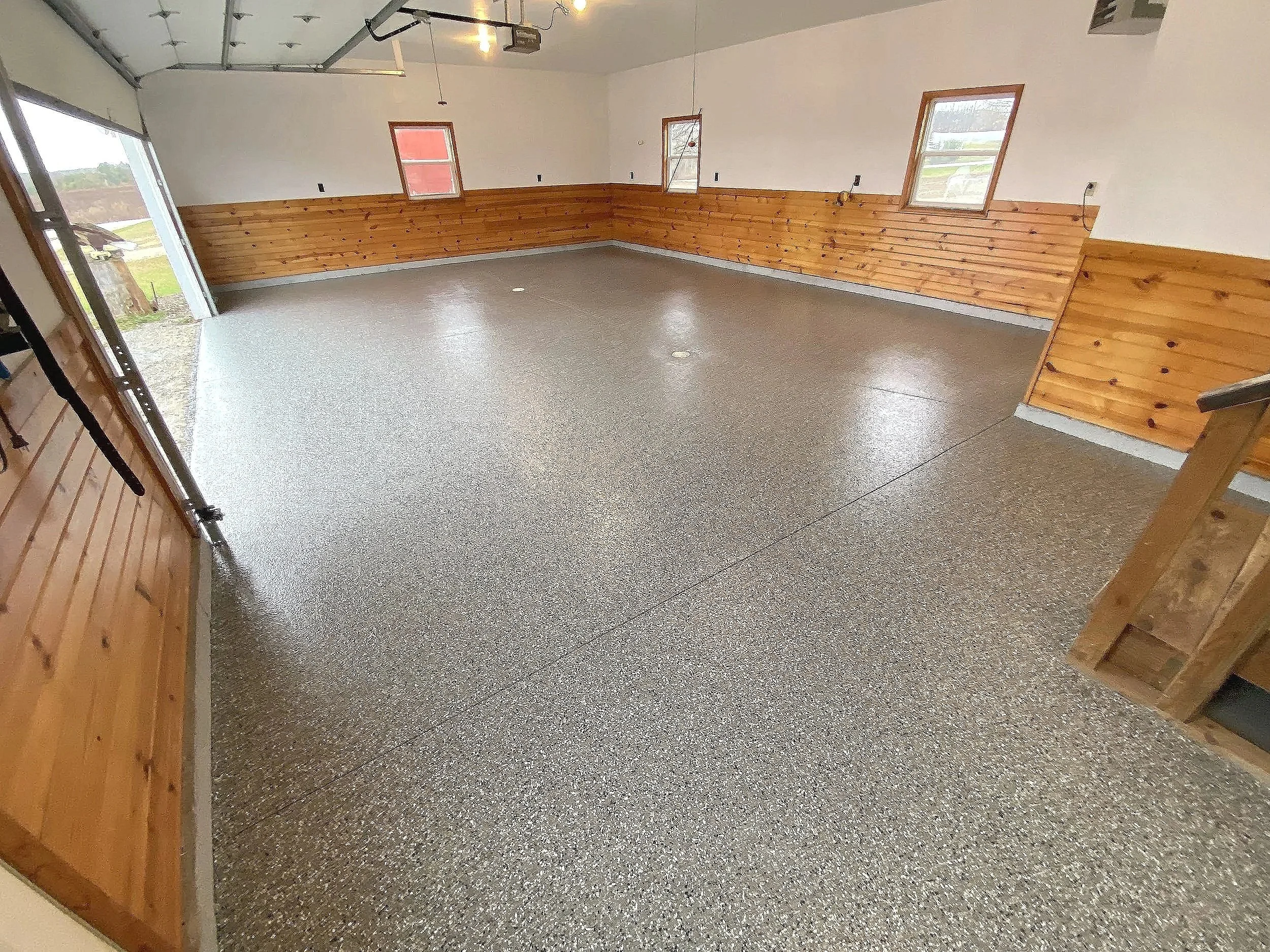 Empty garage with wood-paneled walls, three windows, a door, and a speckled concrete floor.