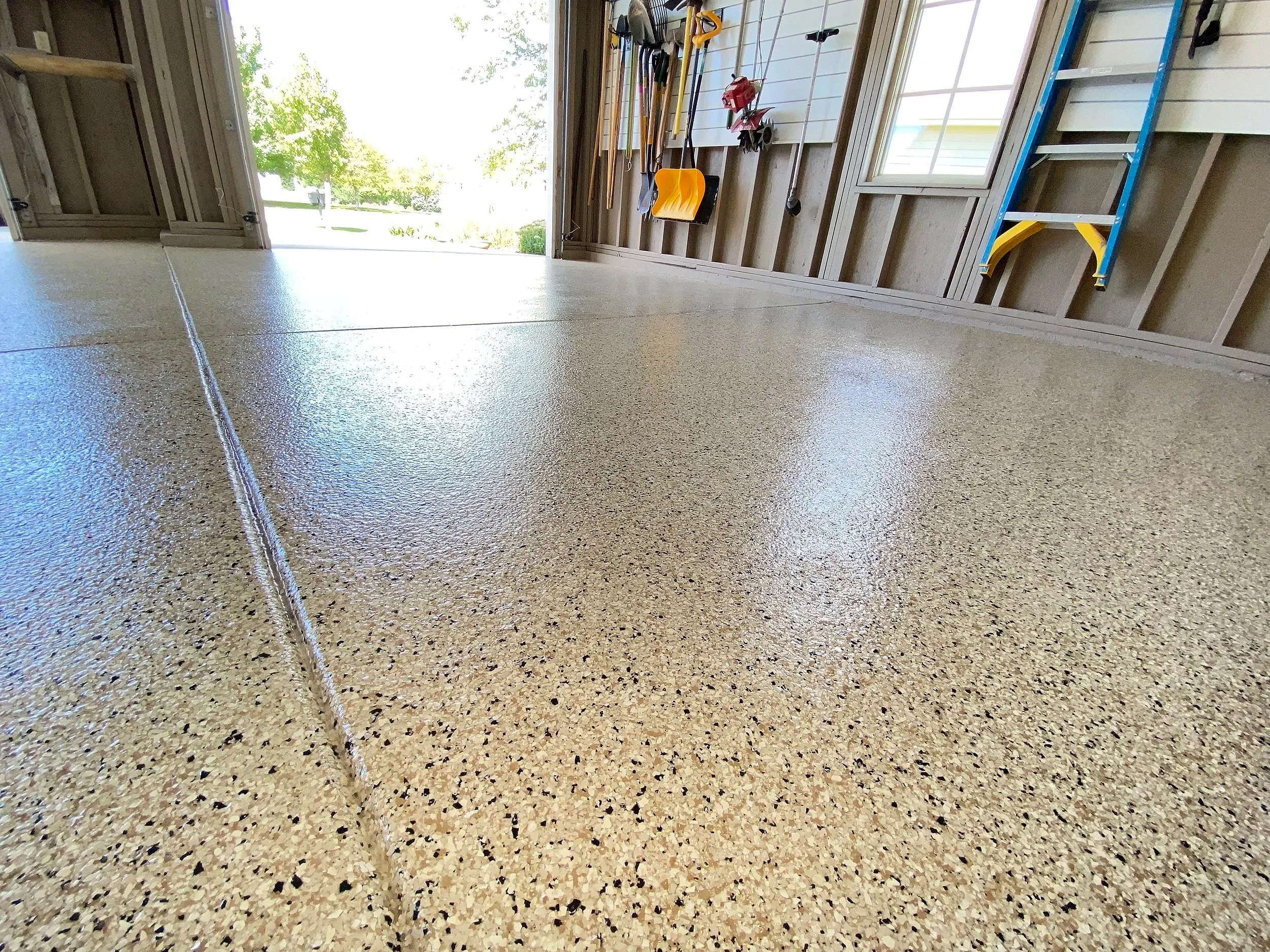 Inside a garage with a polished speckled concrete floor, tools hanging on the wall, and a blue step ladder on the right side, with an open garage door revealing a view of trees and a sunny yard outside.