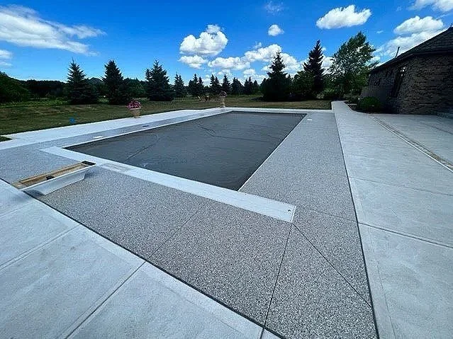Empty backyard swimming pool with cover, surrounded by concrete patio, with trees and blue sky in the background.