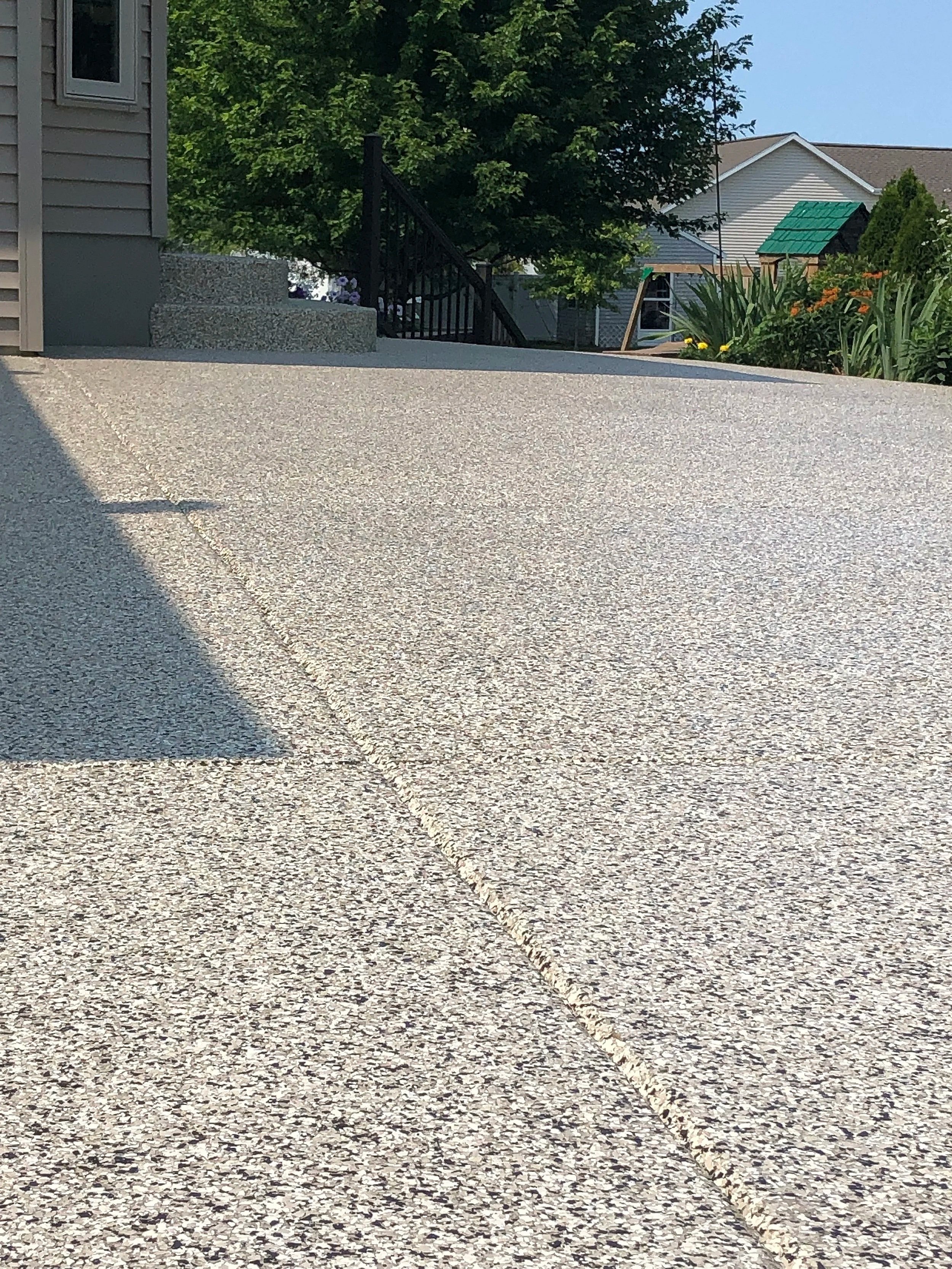 View of a concrete driveway leading to steps near the house with a garden on the right side, trees, and neighboring houses in the background under a clear blue sky.
