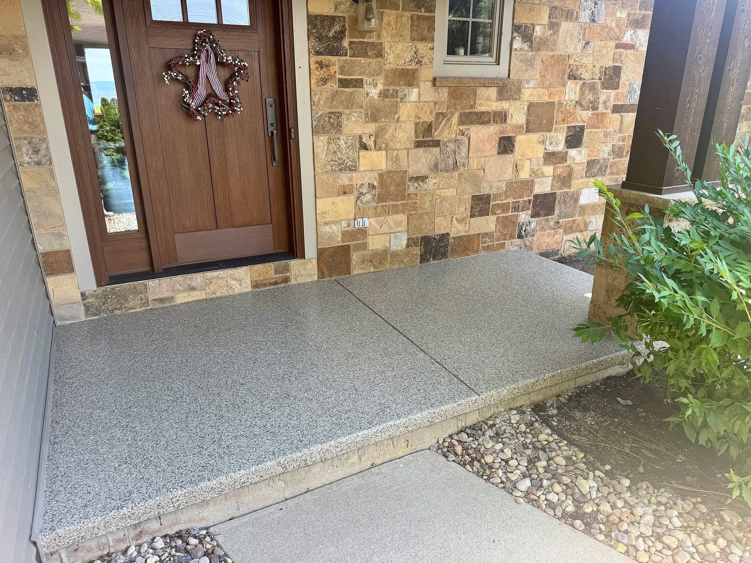 Front porch of a house with stone walls, a wooden door decorated with a holiday wreath, and a concrete step leading to the entrance.