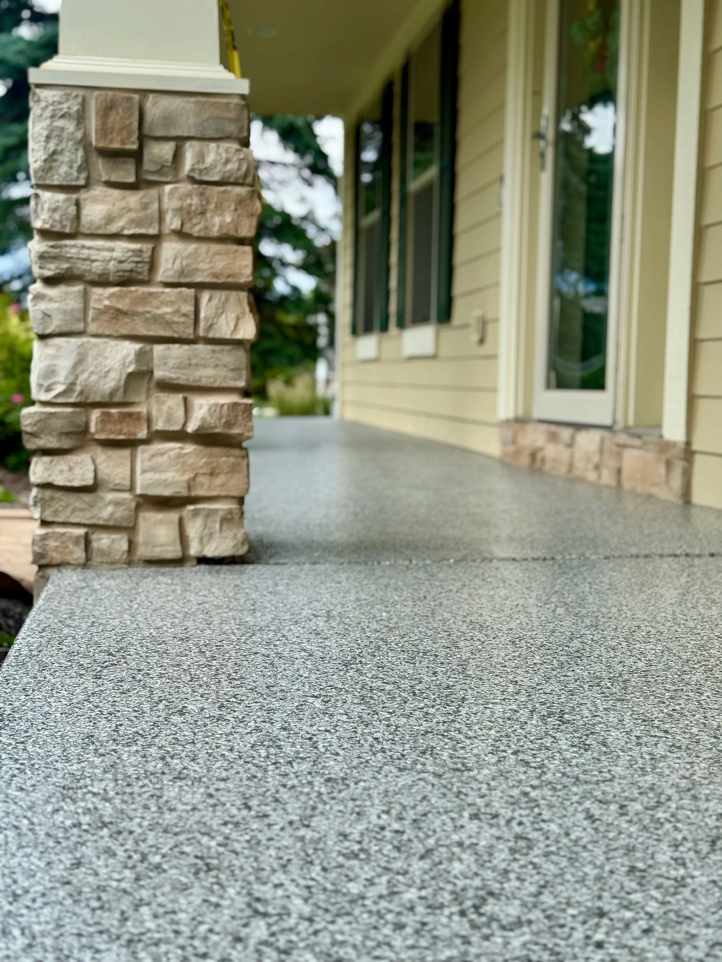Close-up view of a speckled gray porch floor with a stone pillar on the left, part of a house with beige siding, a glass door, and a window in the background.
