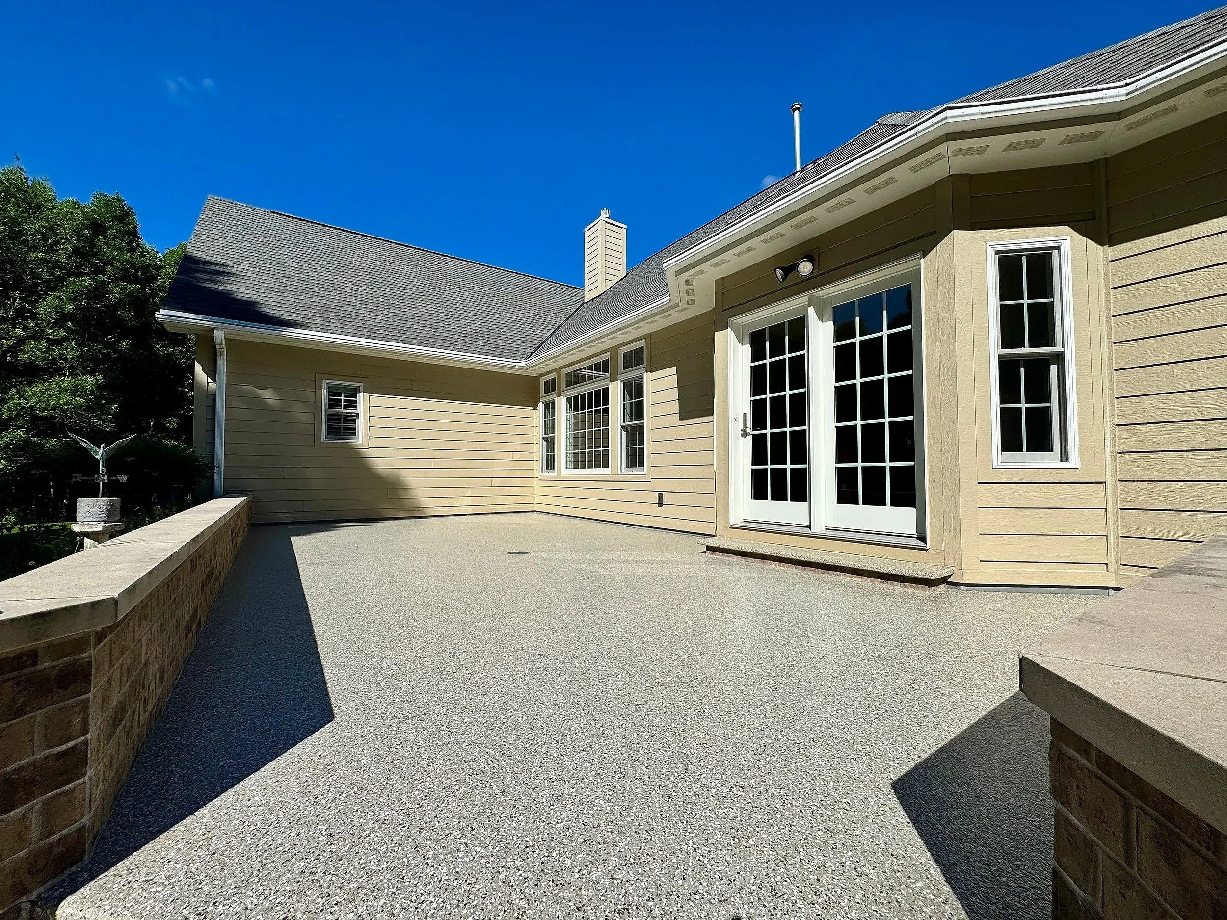 A backyard with a concrete patio, yellow house with white trim, three large windows, a sliding glass door, a white chimney, and a sloped roof against a clear blue sky.
