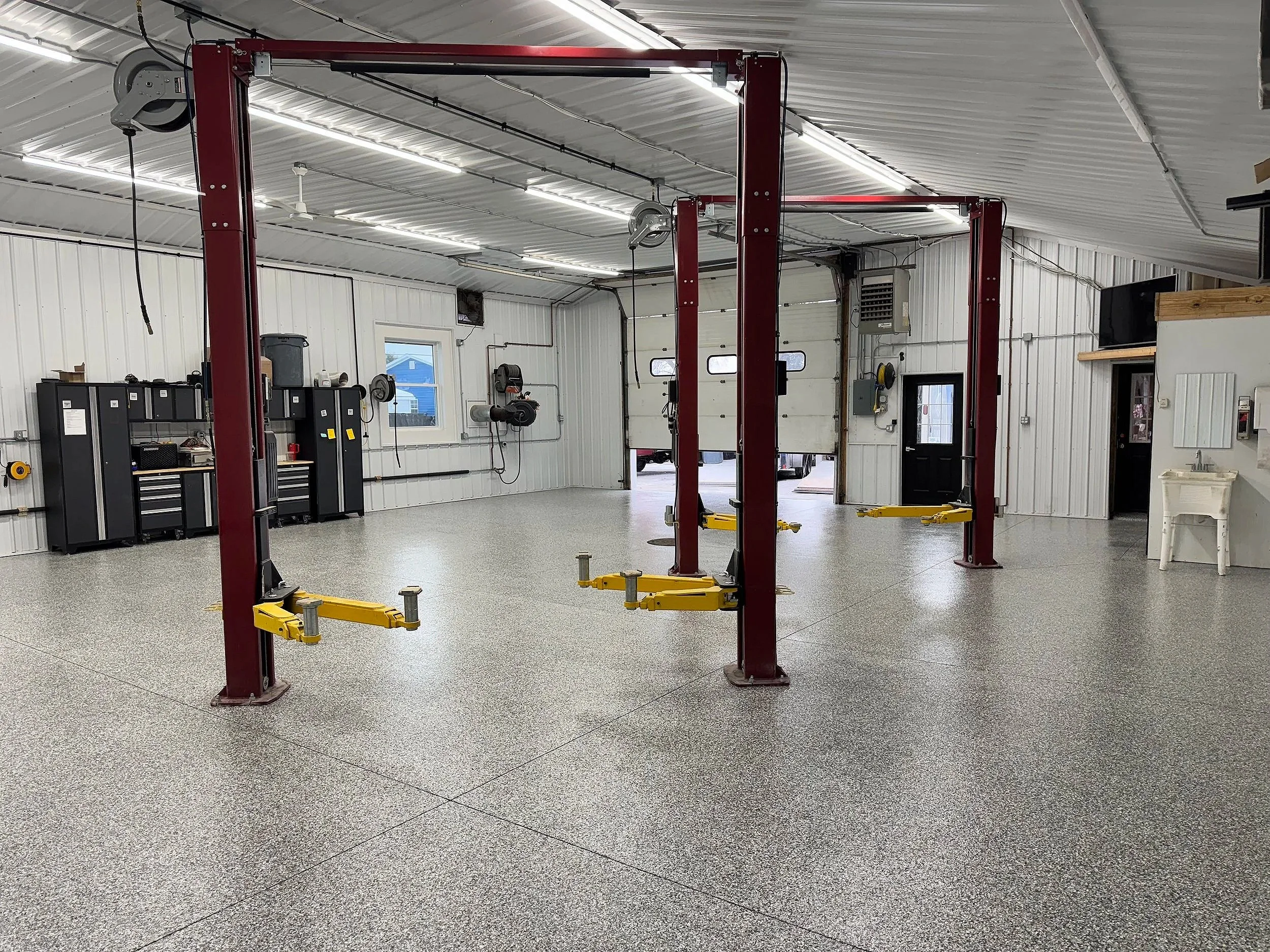 A spacious garage interior with a car lift, tool cabinets, and storage shelves, with white walls, a white ceiling, and fluorescent lighting.