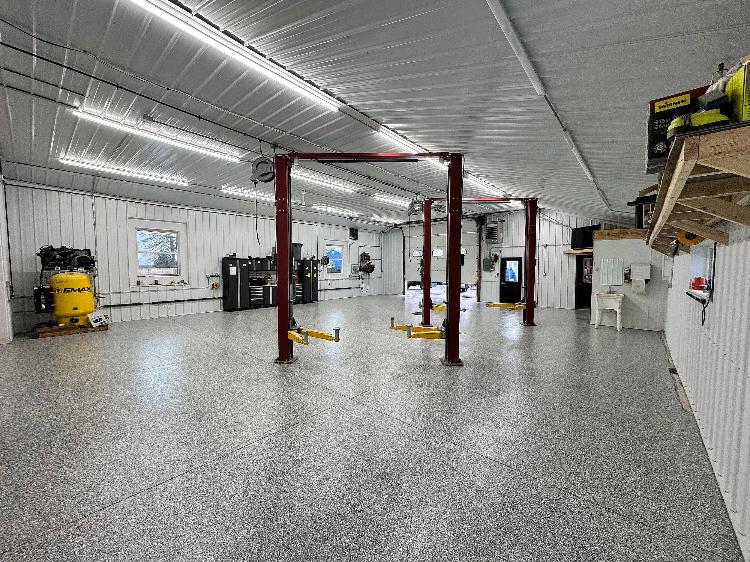 A spacious garage or workshop with white paneled walls and ceiling, and a speckled gray epoxy floor. It contains a red vehicle lift, tool cabinets, a yellow air compressor, and various tools and equipment along the walls. There are small windows and 