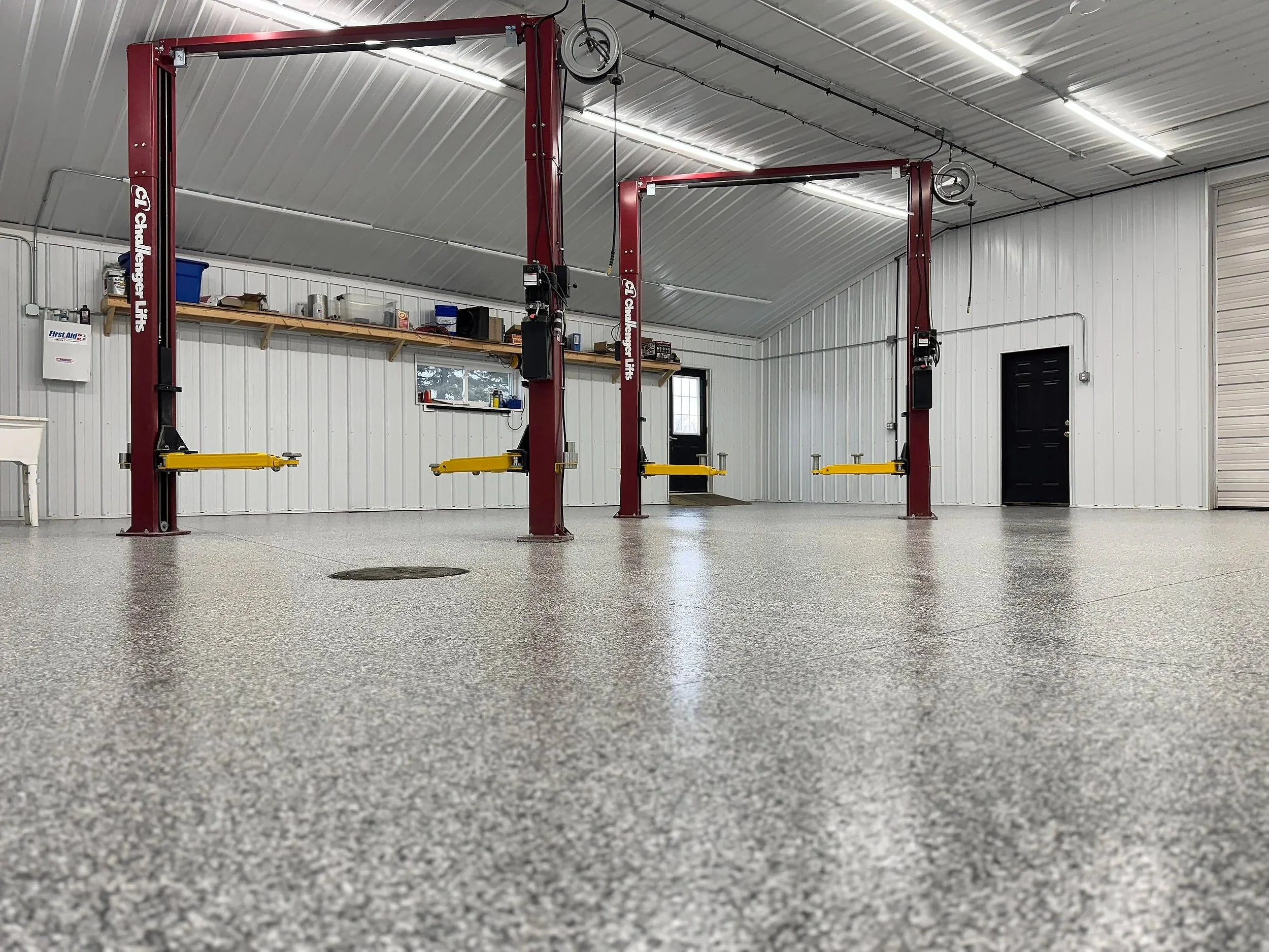 Inside a garage with two car lifts, gray speckled floor, white panel walls, and a metal ceiling with bright lights.