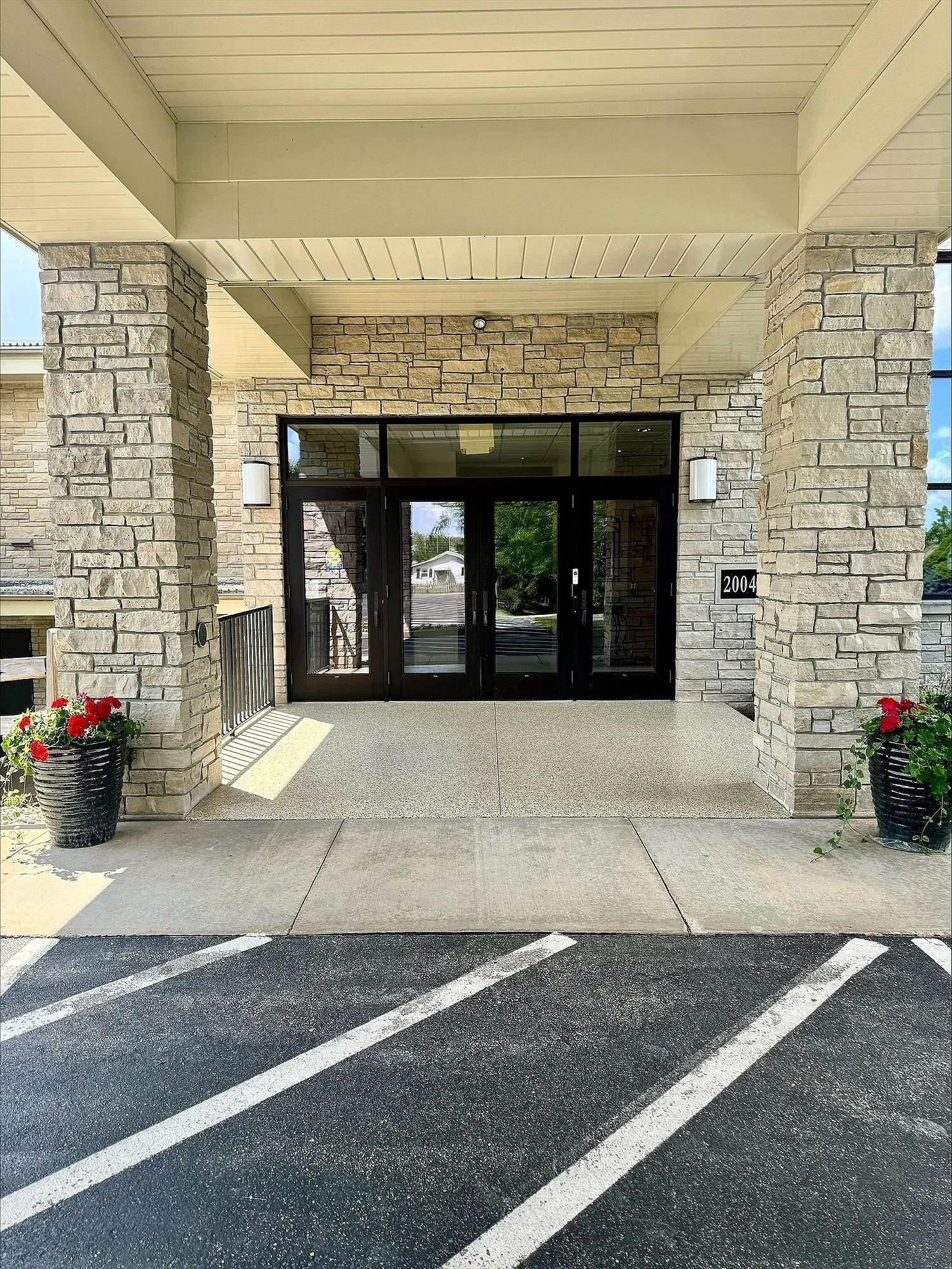 The entrance of a building with stone pillars, glass doors, and potted flowers on each side, with a parking lot in front marked with white lines.