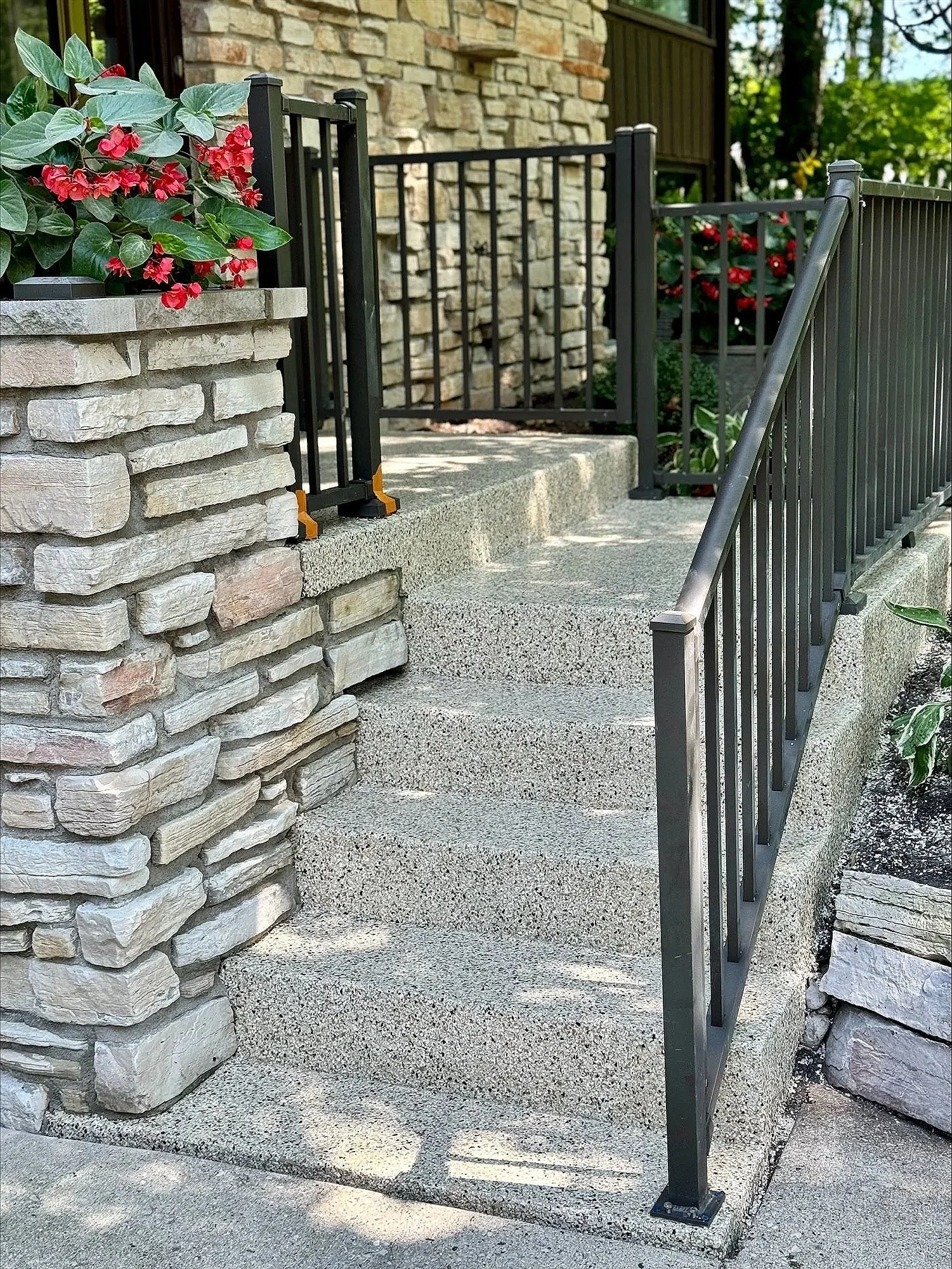 A small black gate with yellow accents installed at the top of stone steps, flanked by stone walls and black metal railing, with flowering plants and trees in the background.