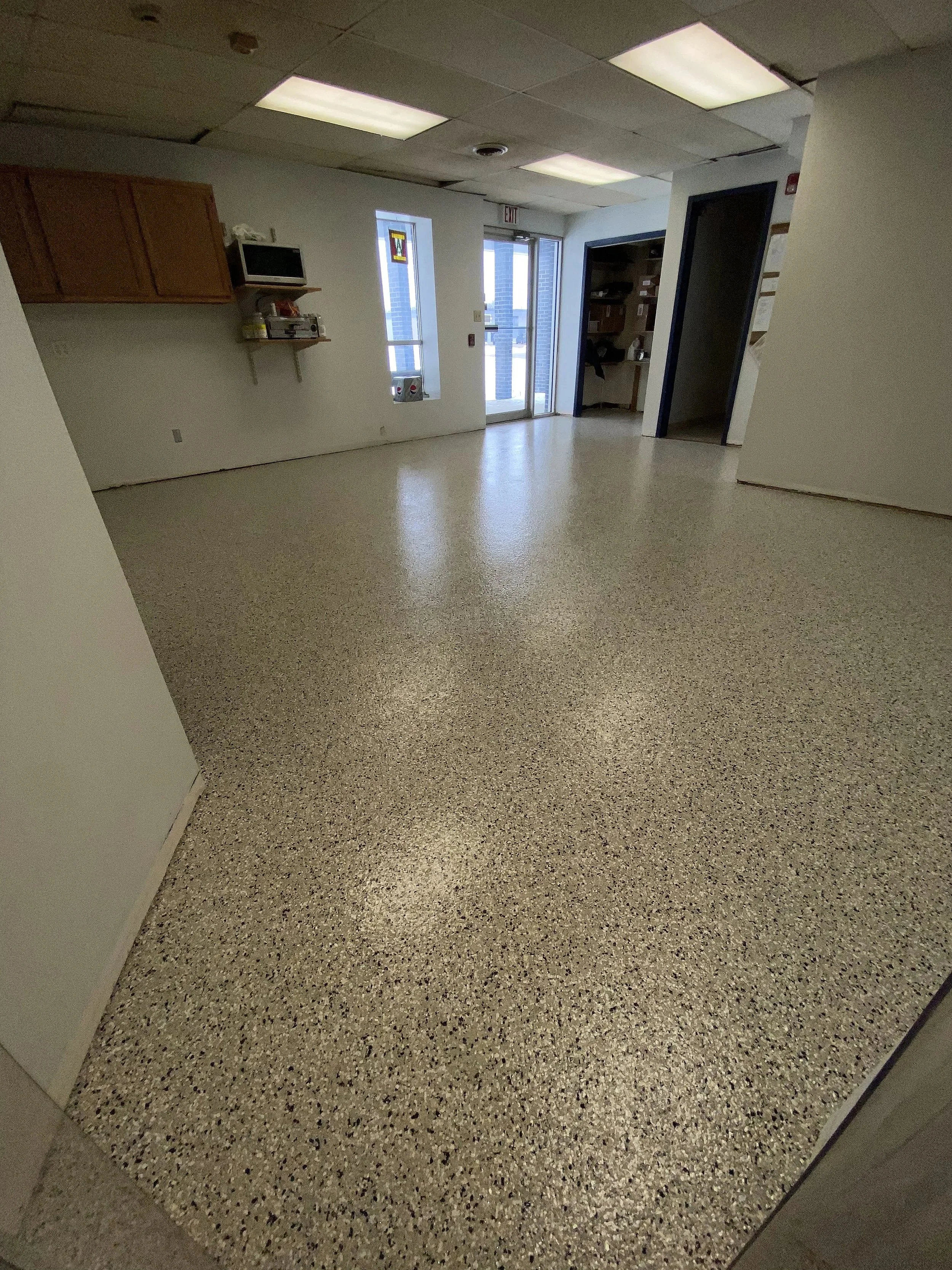 Empty interior room with speckled tile flooring, white walls, ceiling tiles, and fluorescent lighting, with a door and window on the far wall.