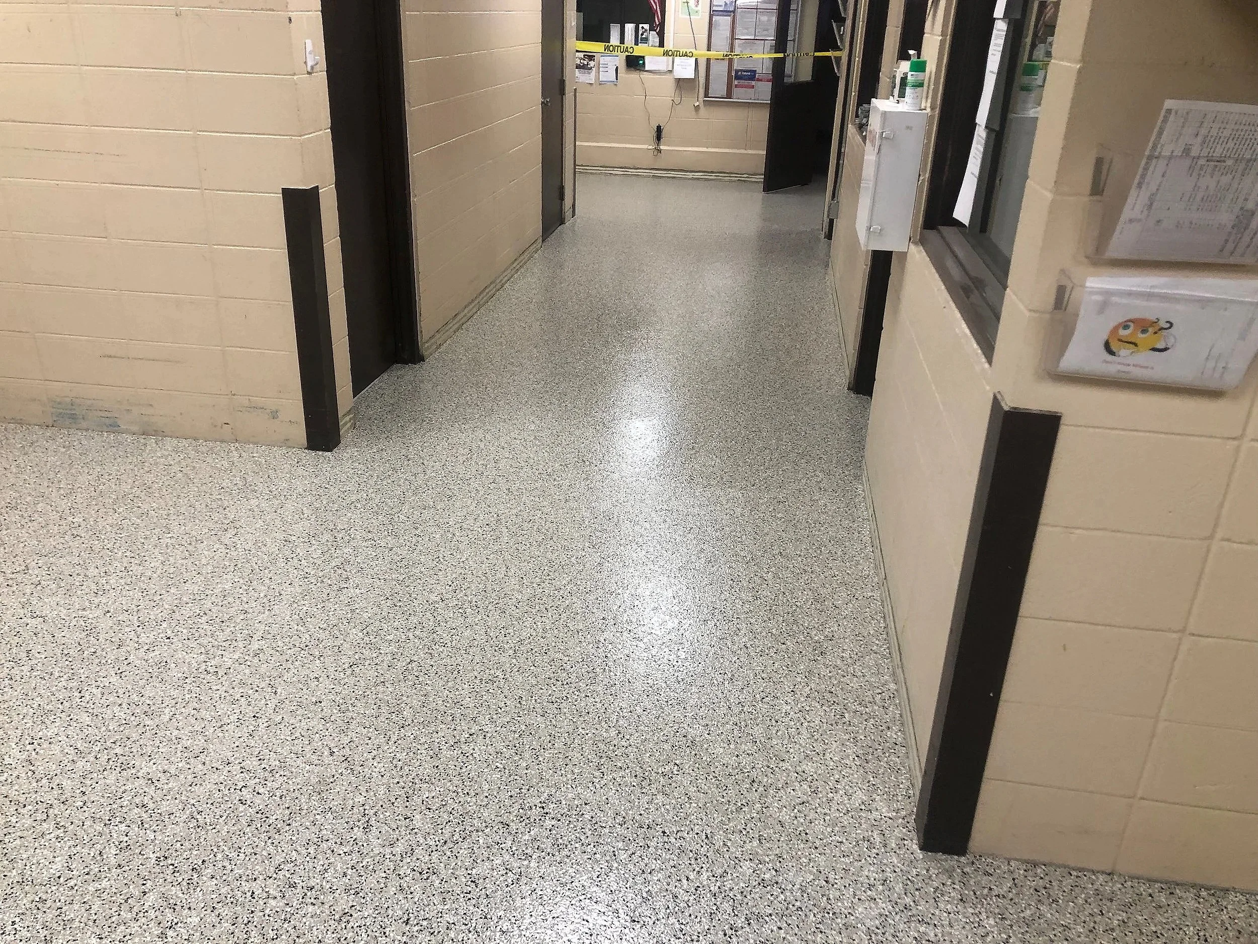 Image of a hallway in a building with beige brick walls, gray speckled flooring, and partial views of doorways. Caution tape is stretched across the hallway, possibly indicating a restricted or closed area. On the right, a window and bulletin board a