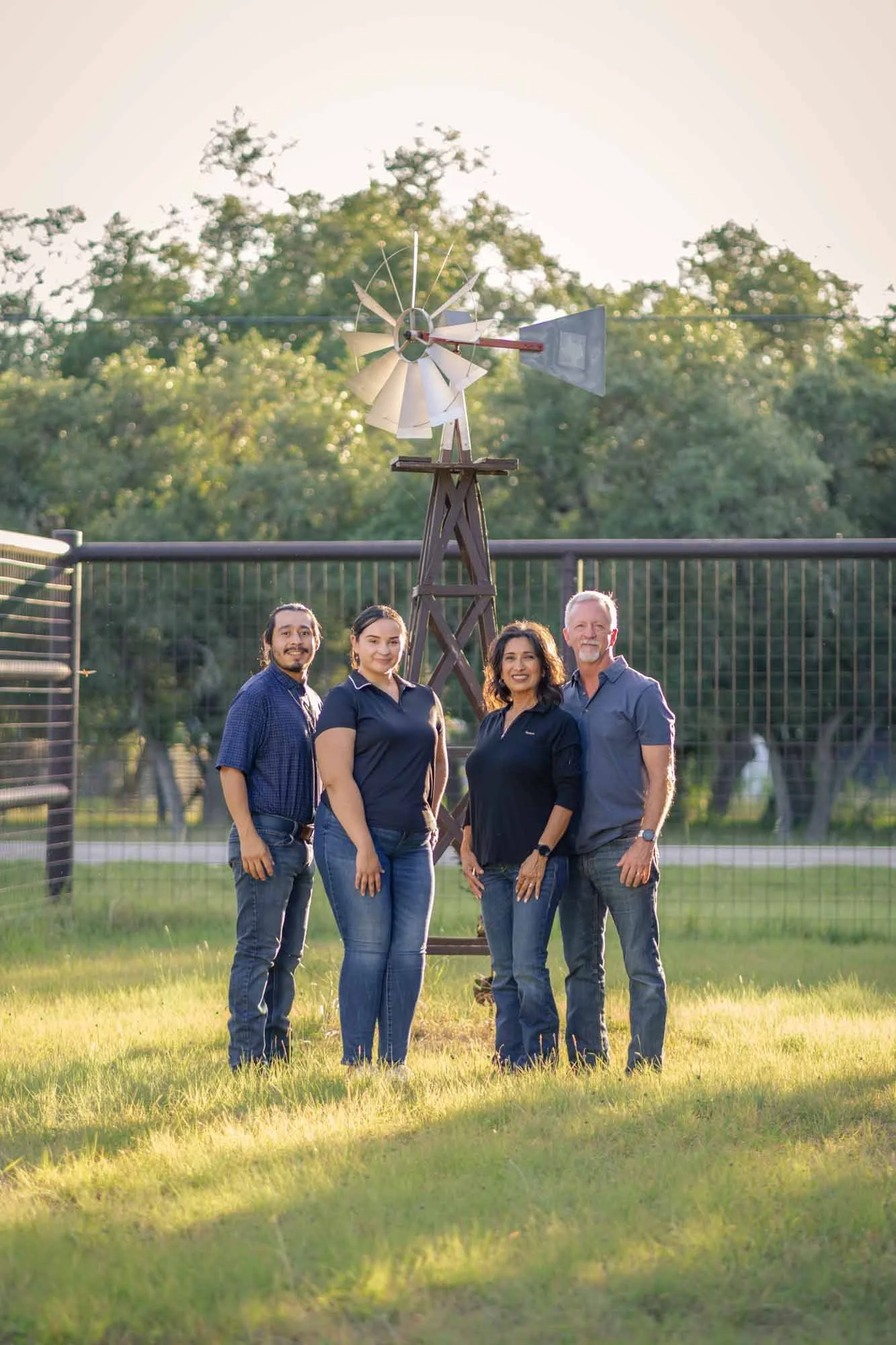 Group of four people standing outdoors in front of a wind turbine model with trees and greenery in the background.