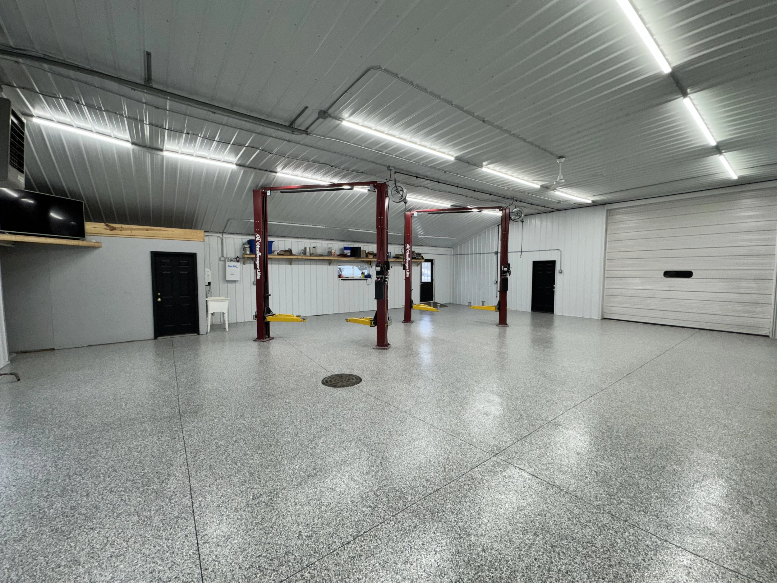 A spacious garage with a metal ceiling, bright LED lights, and two red hydraulic car lifts. The floor is polished concrete and there is a black door and mesh window on the back wall.
