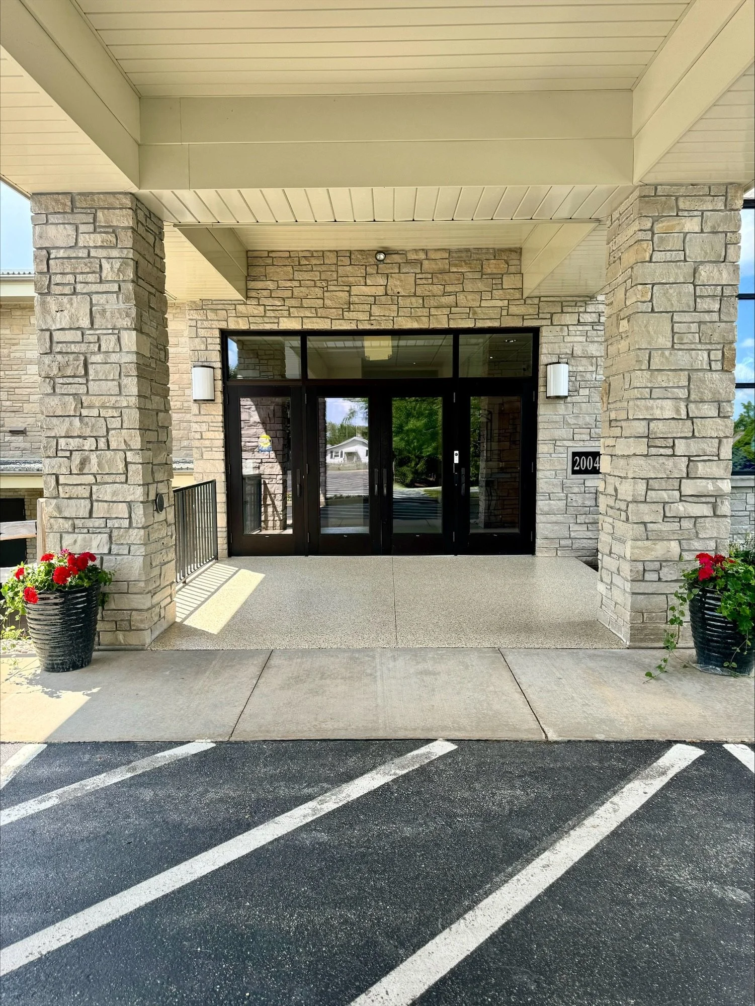 The entrance of a building with stone columns and a glass door, flanked by two planters with red flowers. In front, a parking area with white striped lines on dark asphalt.
