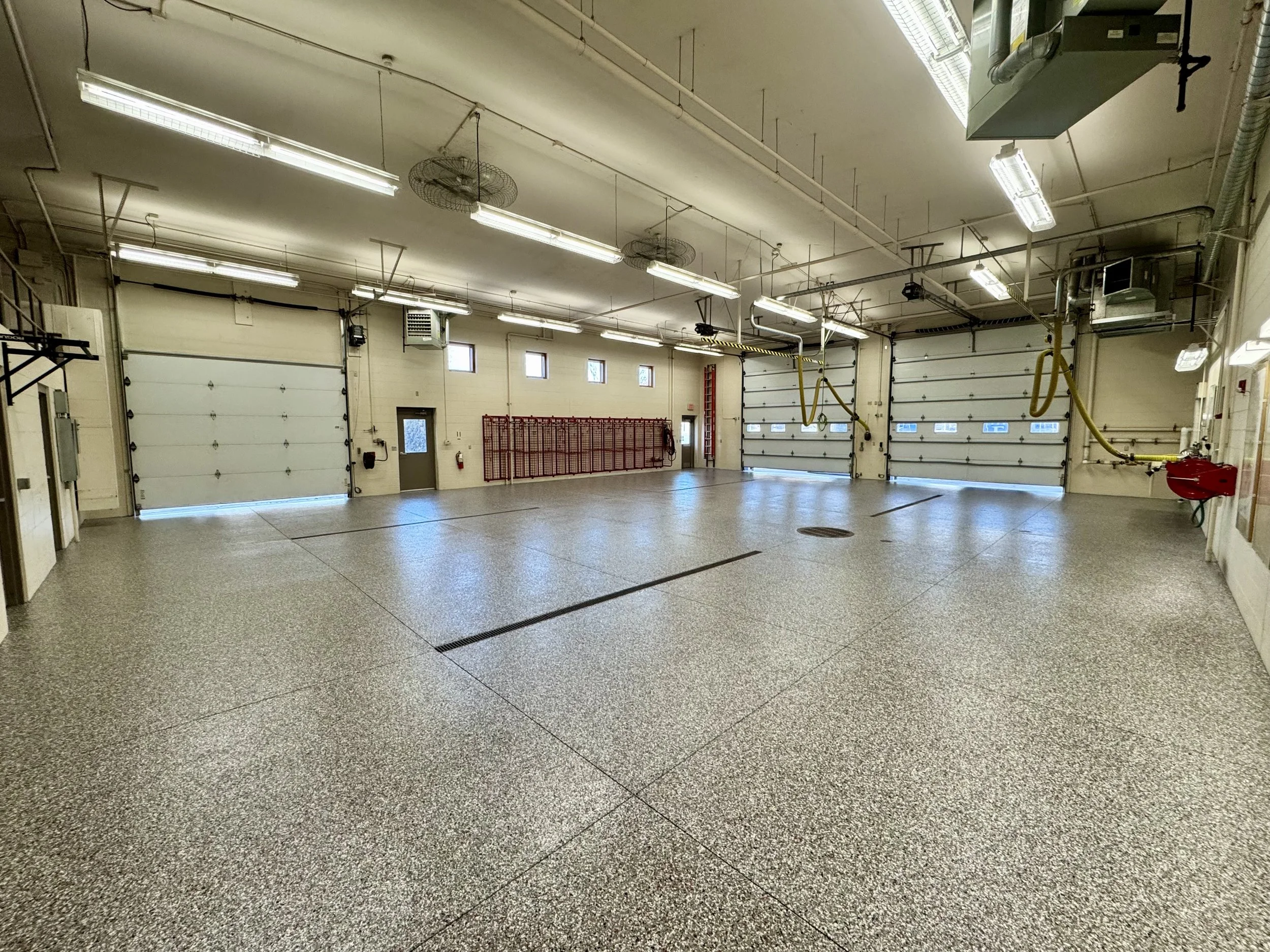 Interior of a garage with three white roll-up doors, high ceilings, fluorescent lighting, ventilation fans, and safety equipment.