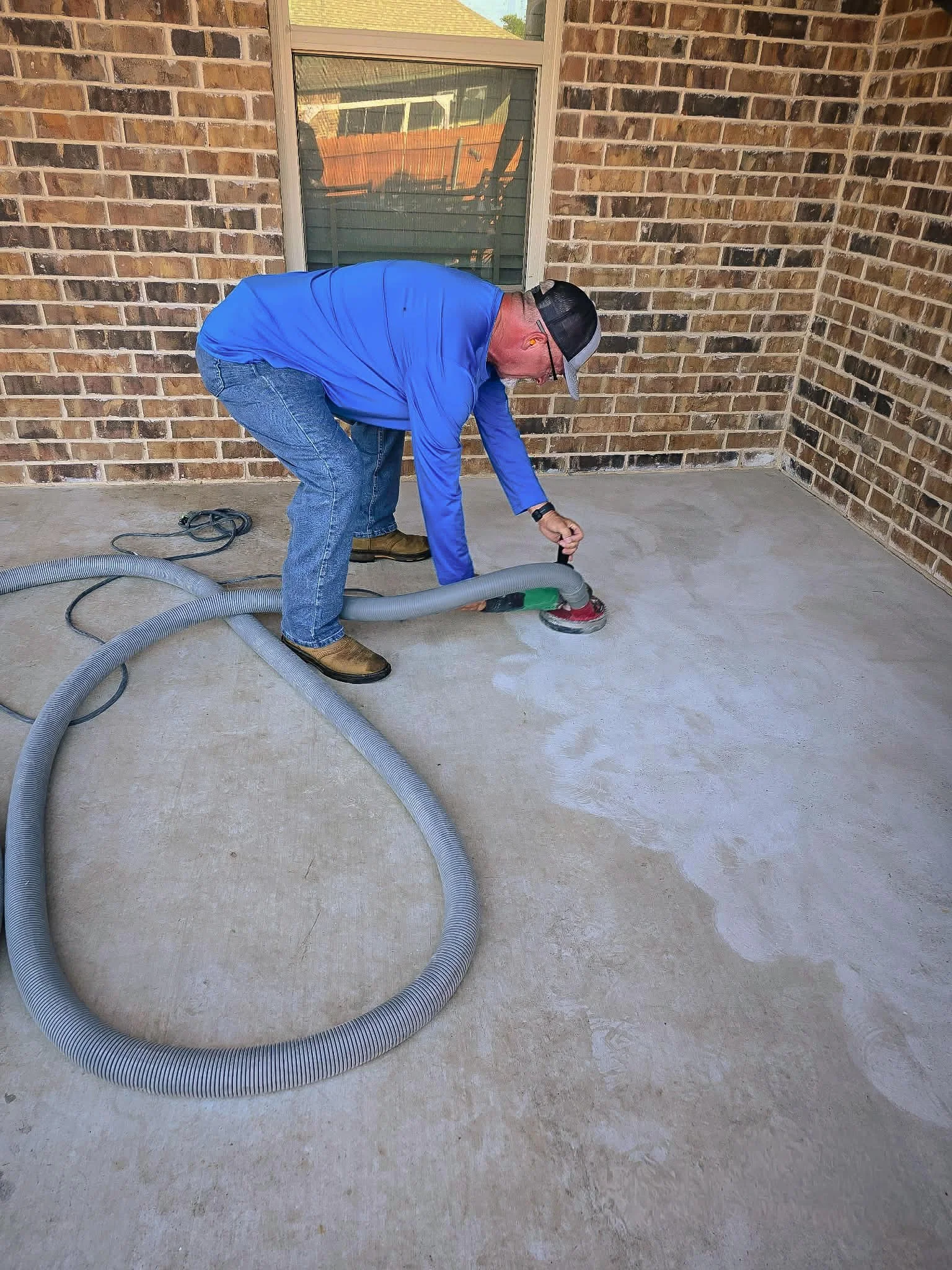 A man in a blue shirt and jeans is vacuuming a concrete porch outside a brick house, with a window and a screened door in the background.