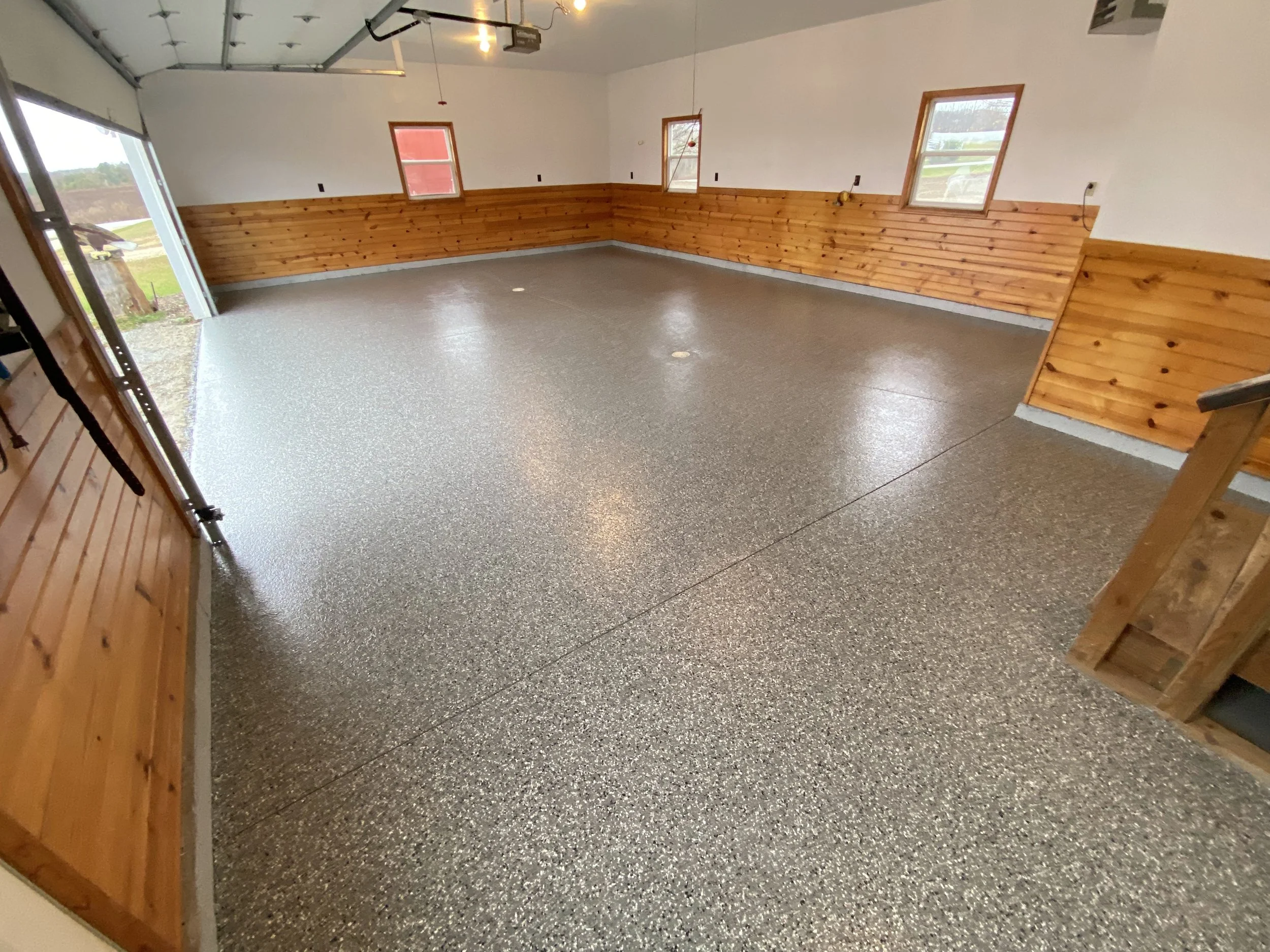 Empty garage with a speckled floor, wooden wainscoting on the walls, five windows, and a partially open garage door.