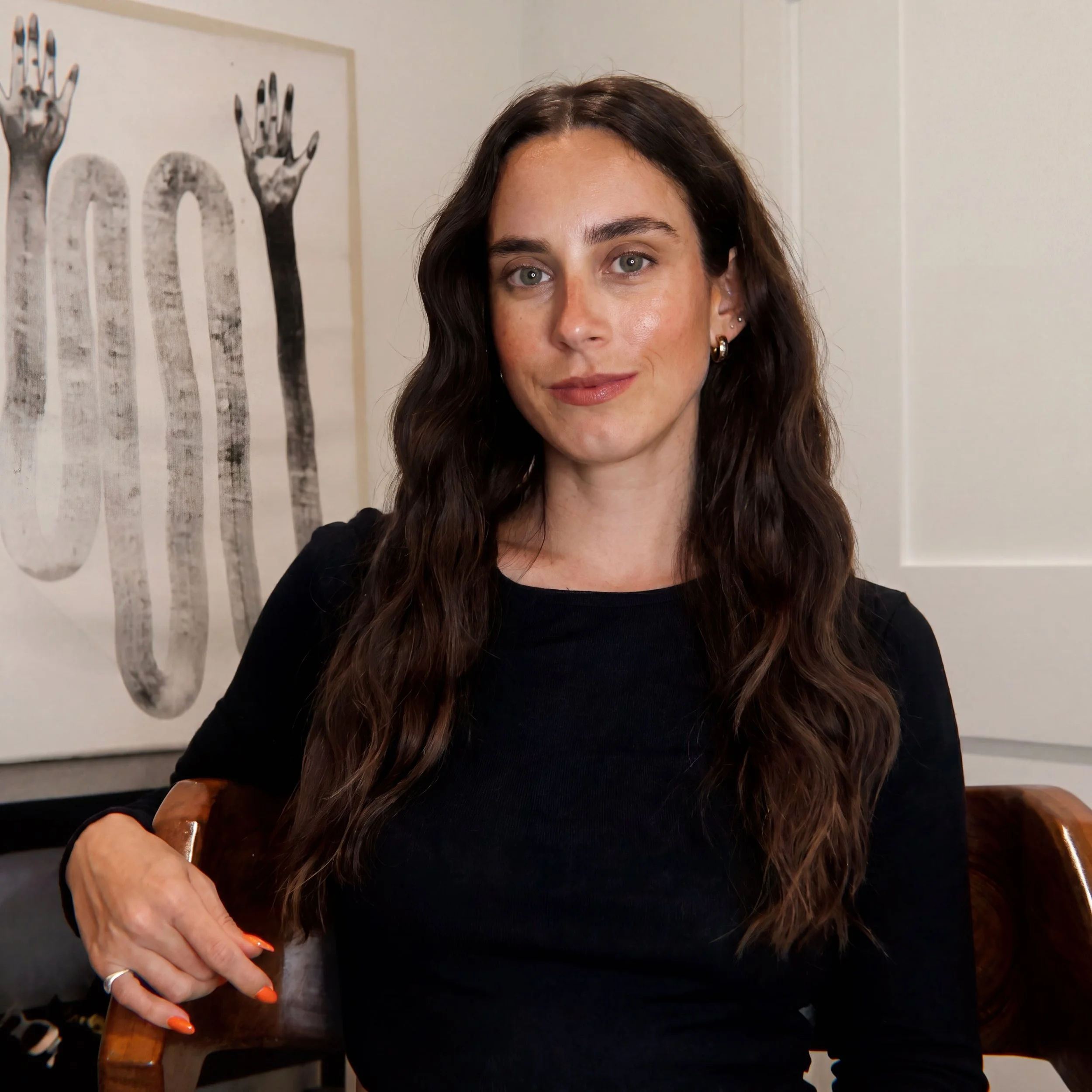 A woman with long brown hair, fair skin, and light-colored eyes, wearing multiple earrings and an orange nail, sitting near a wooden table. Behind her is a wall with a black and white artwork of hands with fingers extended.