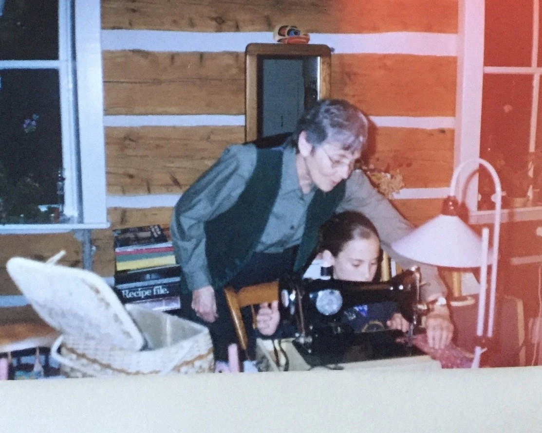 An elderly woman leaning over a young girl, both looking at a sewing machine on a wooden table inside a cozy room with wooden walls. A lamp and books are also on the table.
