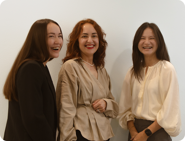 Three women smiling and laughing against a plain white wall, dressed in business casual attire.