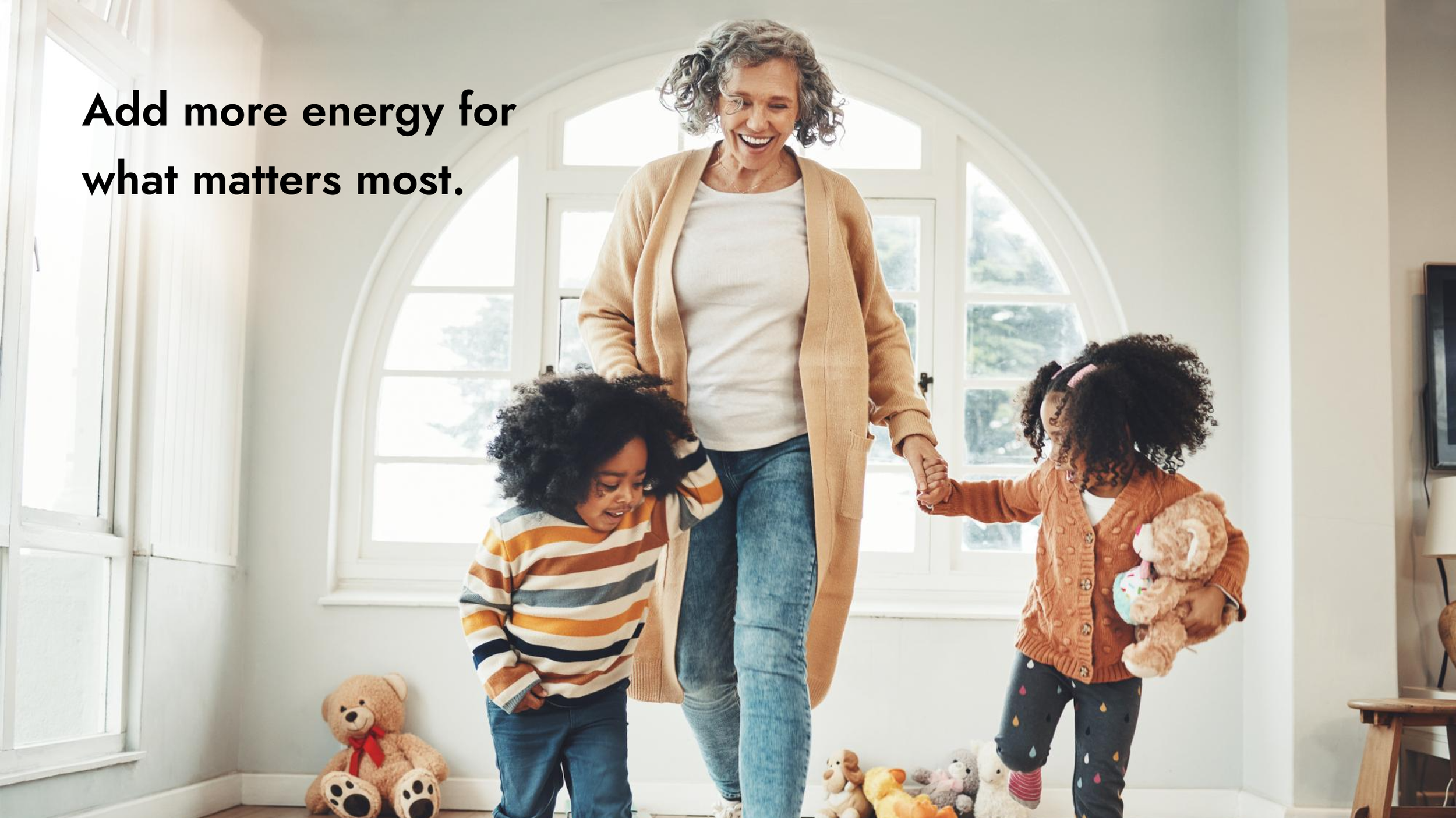 A smiling woman with curly gray hair and glasses, holding hands with two young children in a bright room filled with stuffed animals, including a teddy bear, with large windows and a white wall.