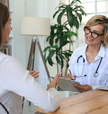 A healthcare professional and a patient having a consultation in a bright room with large windows and a green plant.