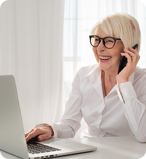 An older woman with blonde hair, glasses, and a white shirt, smiling while talking on a cell phone and using a laptop.