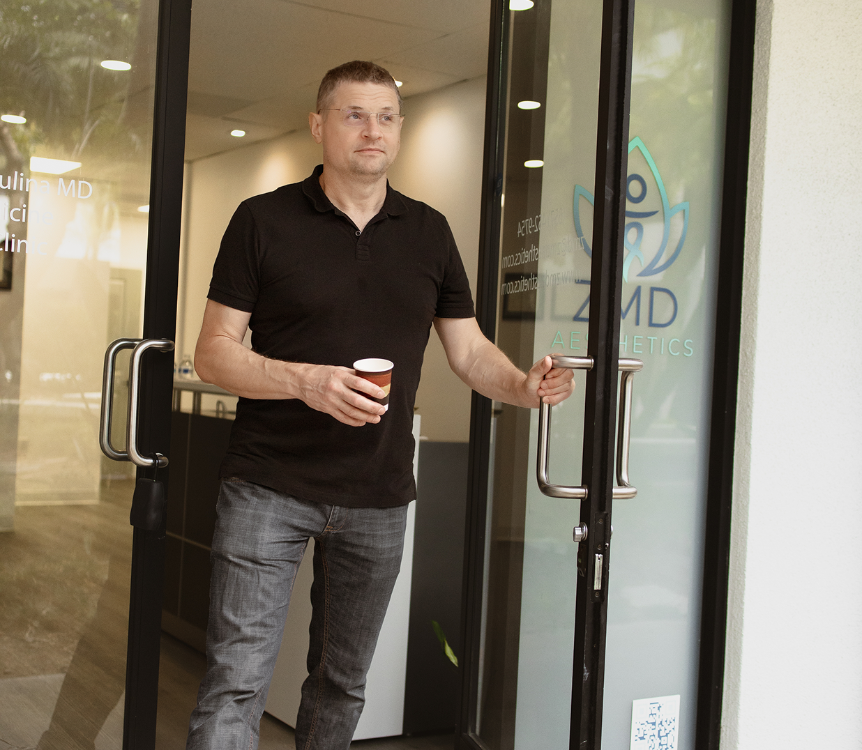 A man stands at the entrance of an aesthetics clinic holding a coffee cup, wearing a black polo shirt and jeans, with glass doors behind him displaying the clinic's logo and name.
