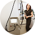 Woman in a black shirt and white pants standing in front of a medical X-ray machine in a clinical setting.