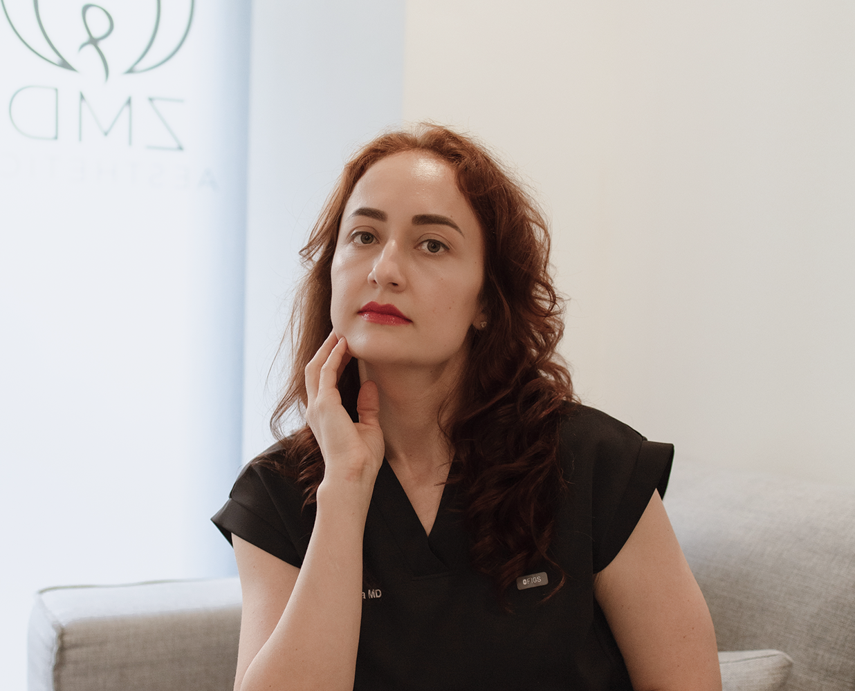 A woman with long, wavy reddish-brown hair, wearing black scrubs, sitting on a gray couch, resting her chin on her hand, looking at the camera in a medical office setting.