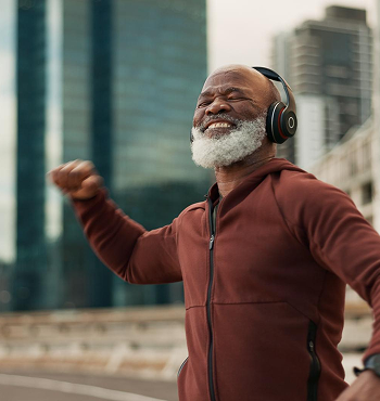 A joyful senior man with white beard and hair wearing headphones, running outdoors in an urban area with glass buildings, smiling with his eyes closed.