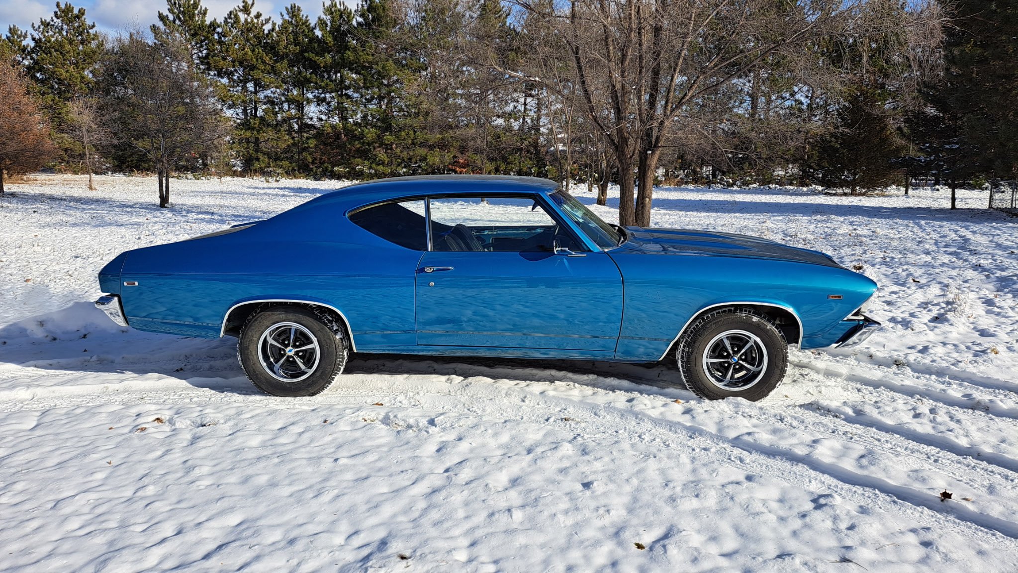 A blue vintage car parked on snow-covered ground with leafless trees and evergreen trees in the background.